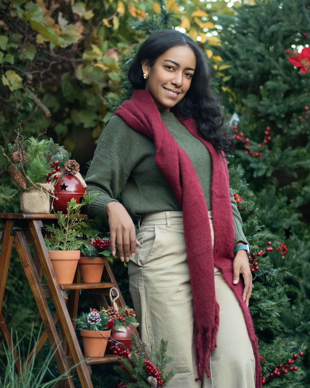 Gardener in olive coat and burgundy scarf arranges potted plants on wooden ladder shelf in lush garden setting.