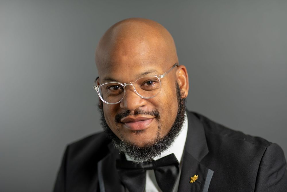Professional portrait of person in black bow tie and glasses against gray background.
