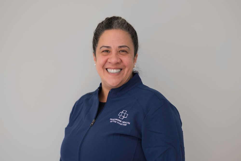 Medical professional in navy quarter-zip uniform smiling against gray background.