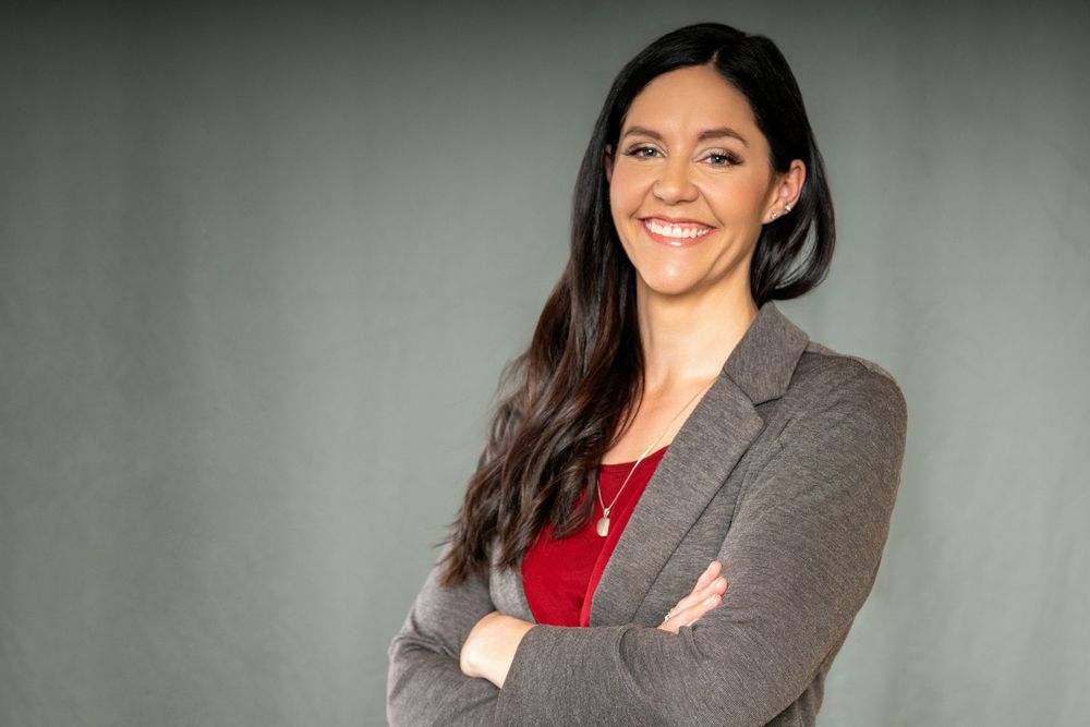 Person in red top and grey blazer poses confidently against neutral backdrop.