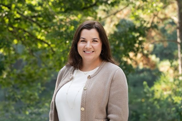 Professional outdoor headshot of a smiling businesswoman wearing a beige cardigan over a white blouse with greenery in the background.