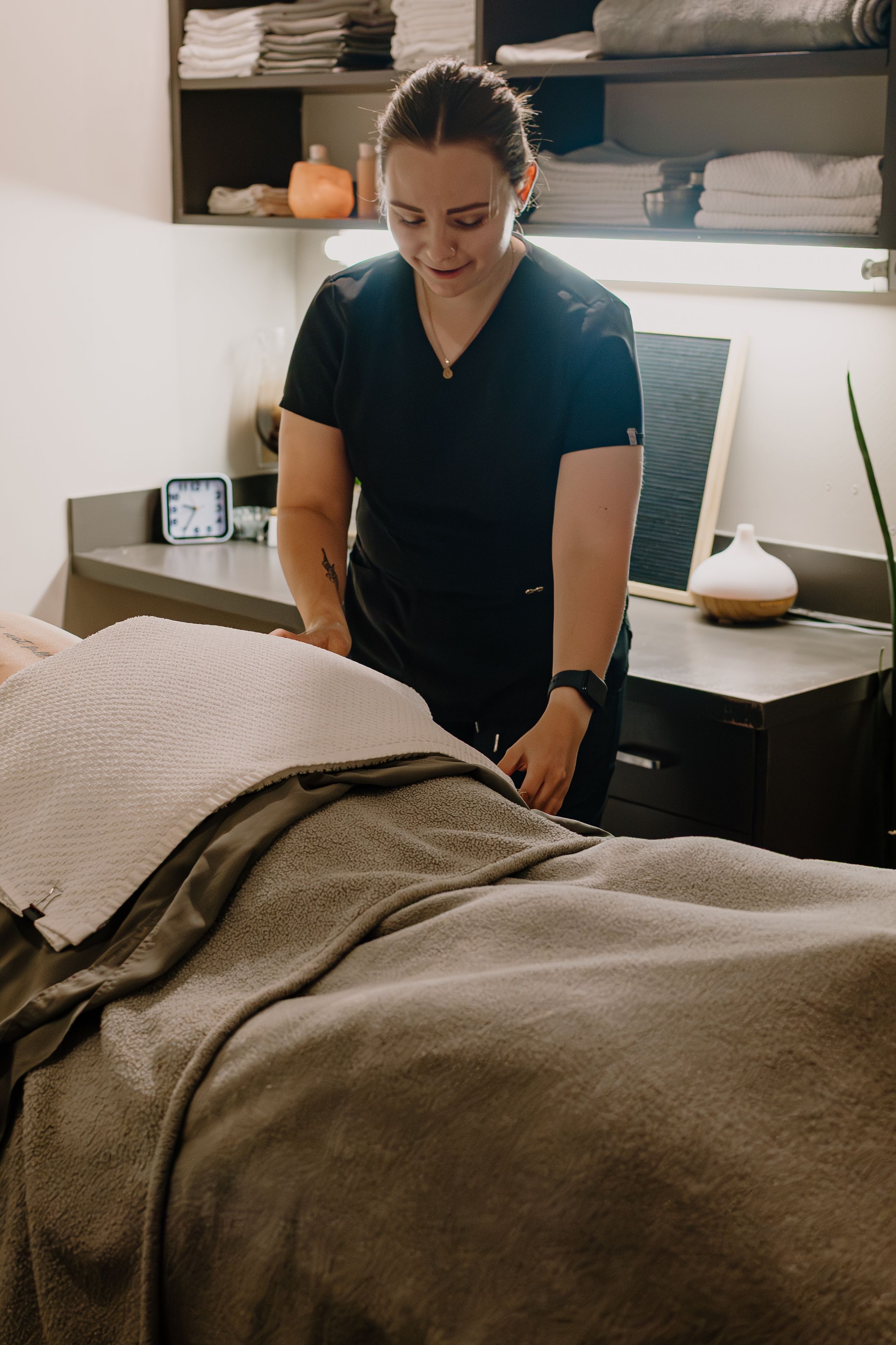 Massage therapist in black uniform prepares treatment table with brown linens in a serene spa setting.