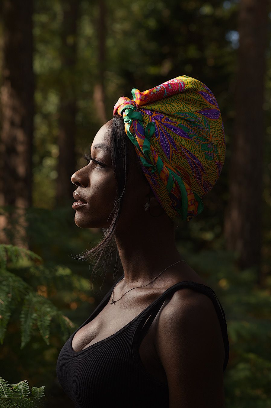 Woman in black tank top and colorful headscarf poses thoughtfully in forest setting during golden hour.