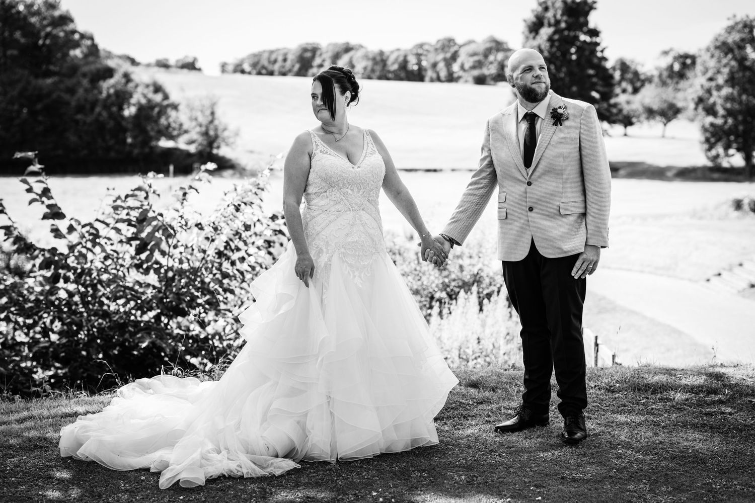 A couple walks along a scenic outdoor path during their wedding photoshoot on a bright sunny day.