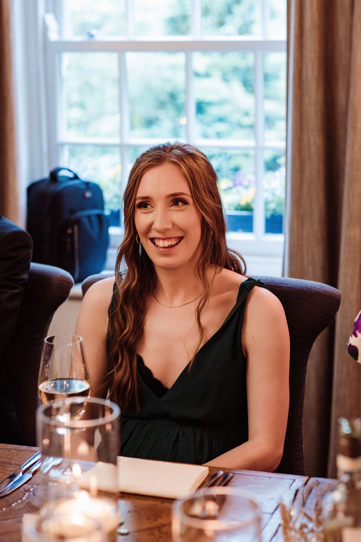 A person in a black sleeveless top smiles warmly while seated at a dining table with glassware.