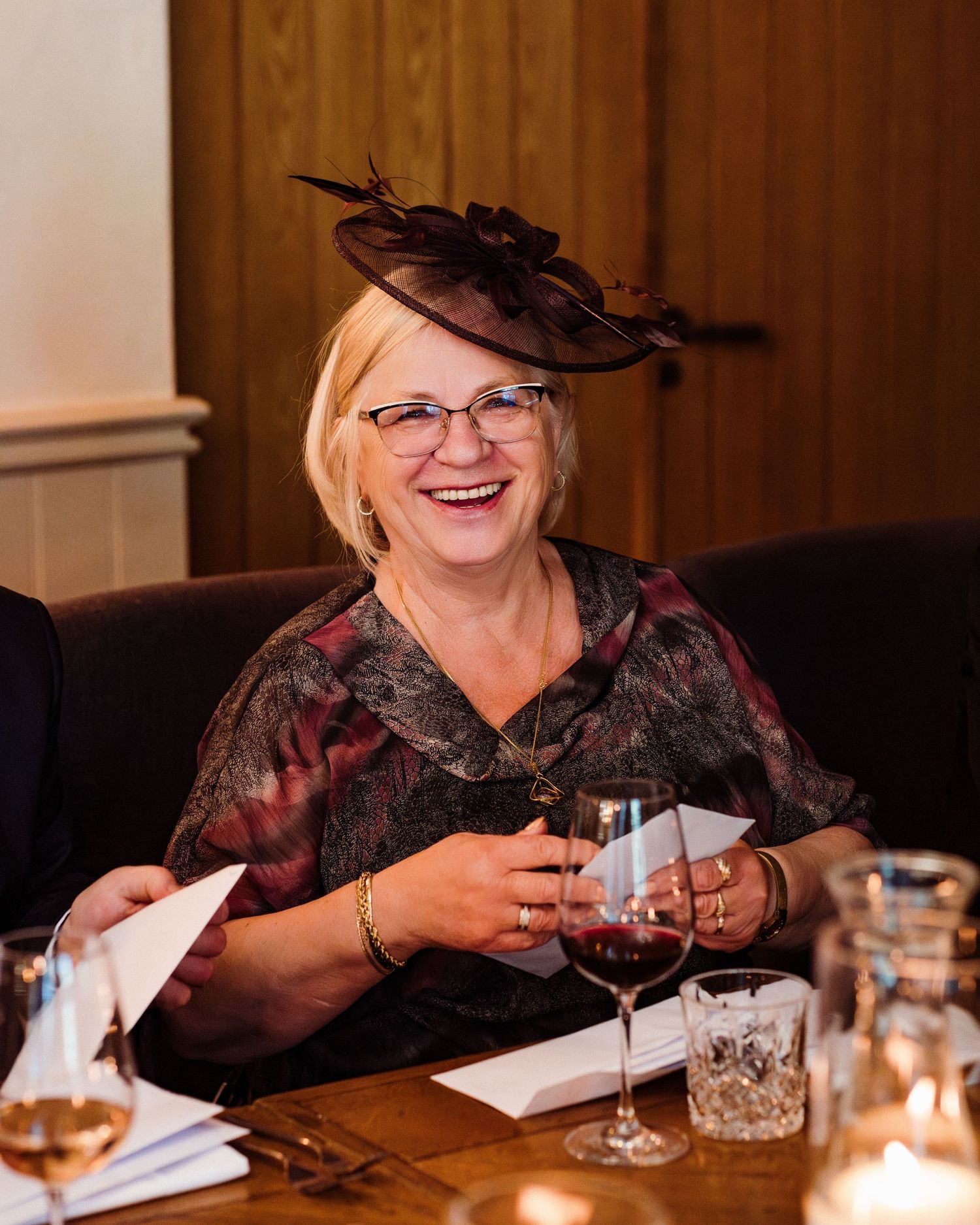 A guest wearing a decorative hat and glasses smiles while holding a wine glass at a formal event.