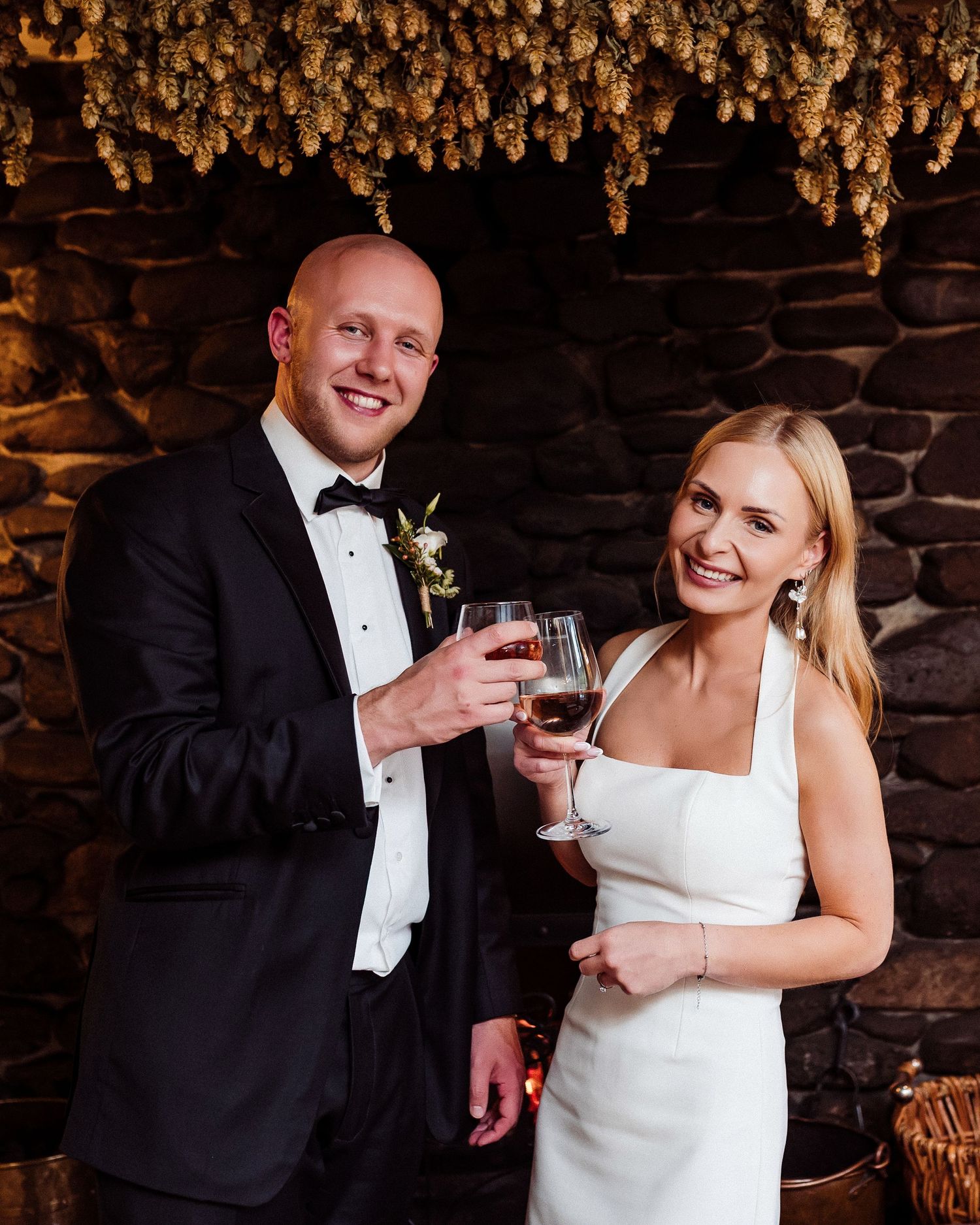 A couple in formal wedding attire toasting with wine glasses in front of a rustic stone wall.