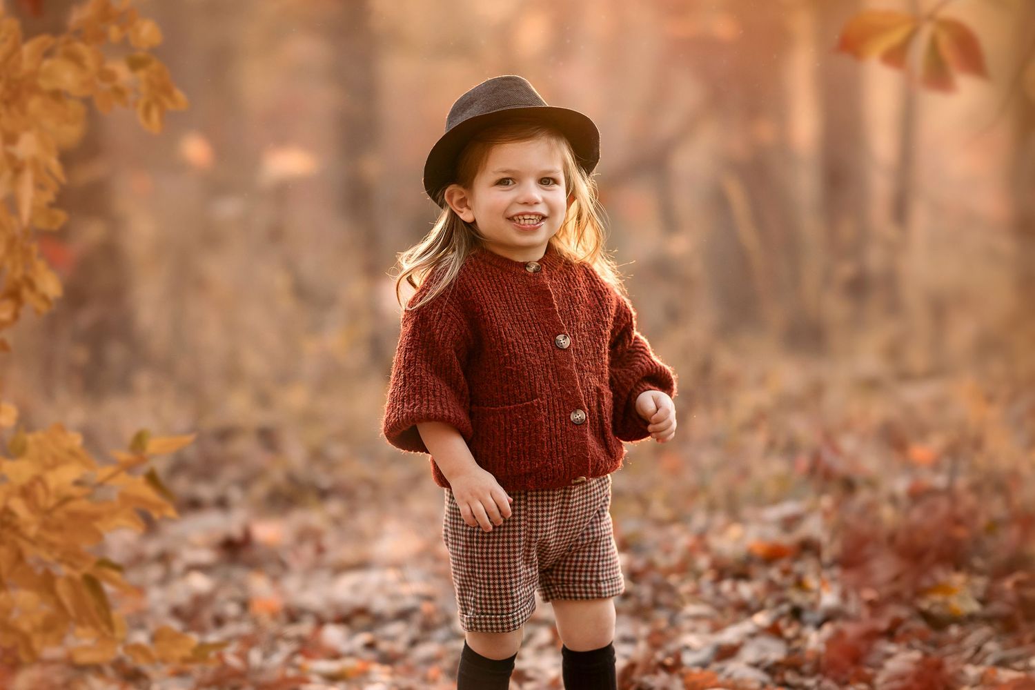 A small child plays in fallen autumn leaves wearing a brown sweater and hat during a golden sunset.