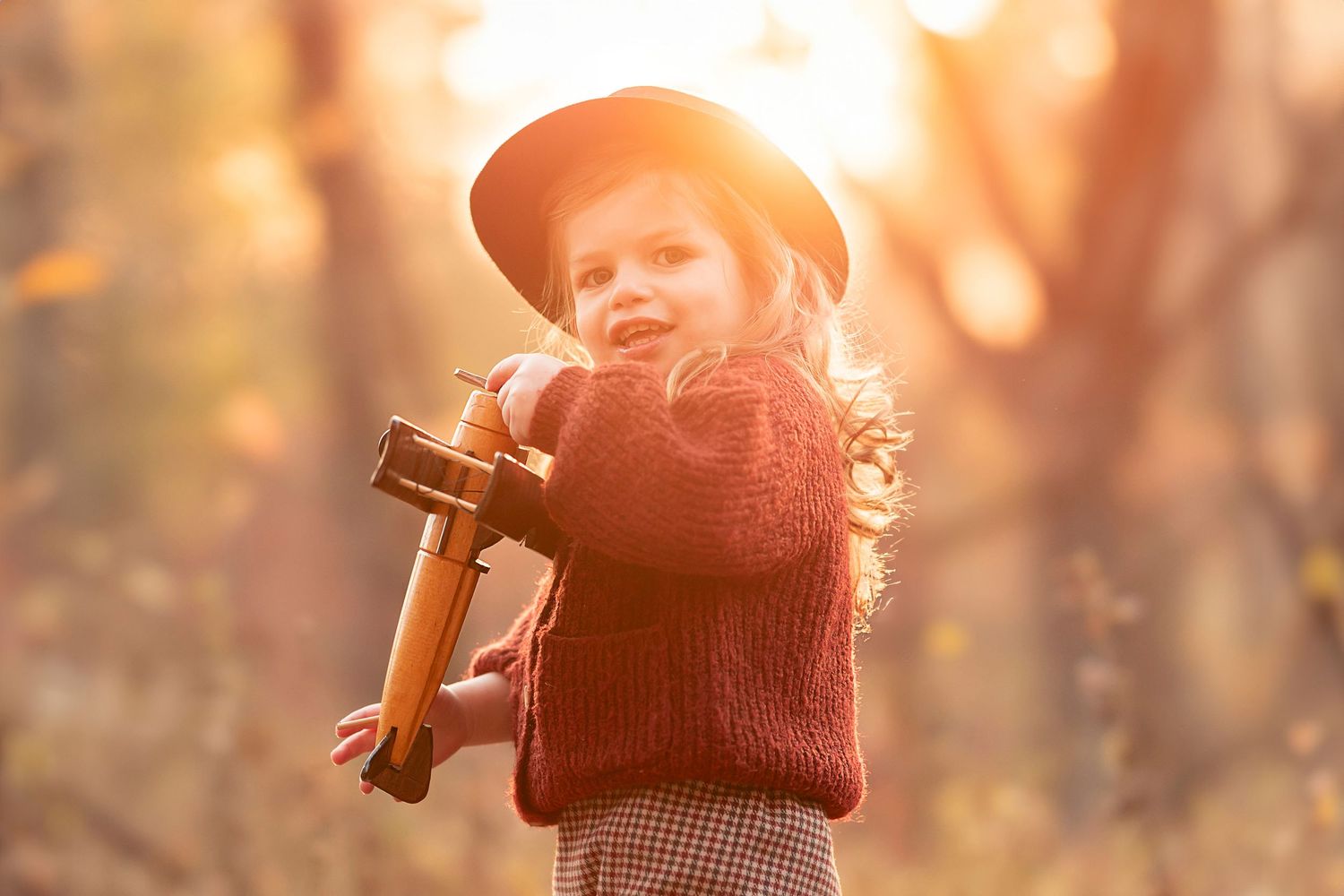 A person in a knit beanie holds a vintage camera in a sunlit forest setting with warm golden bokeh lighting.