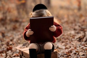 Small child in a red sweater sits on wooden crate reading a book among fallen autumn leaves.