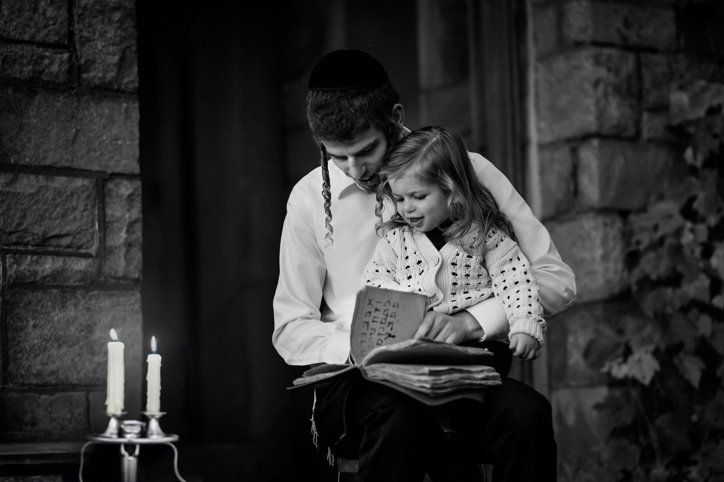 A couple in vintage attire reads together by candlelight in black and white photograph.