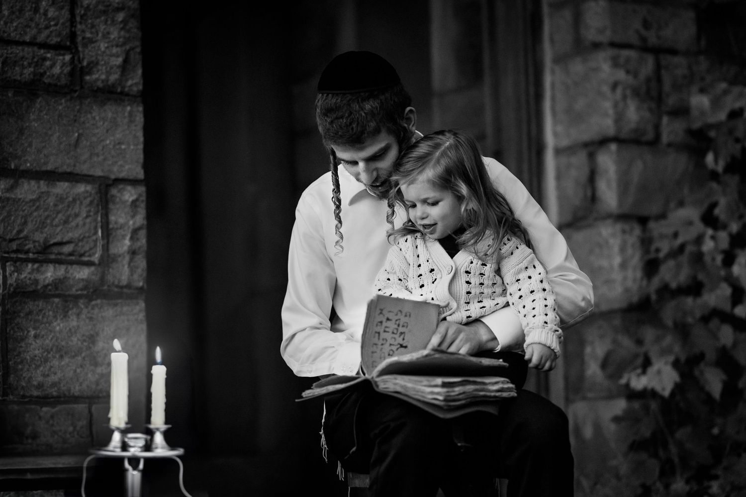 A couple in vintage attire reads together by candlelight in black and white photograph.