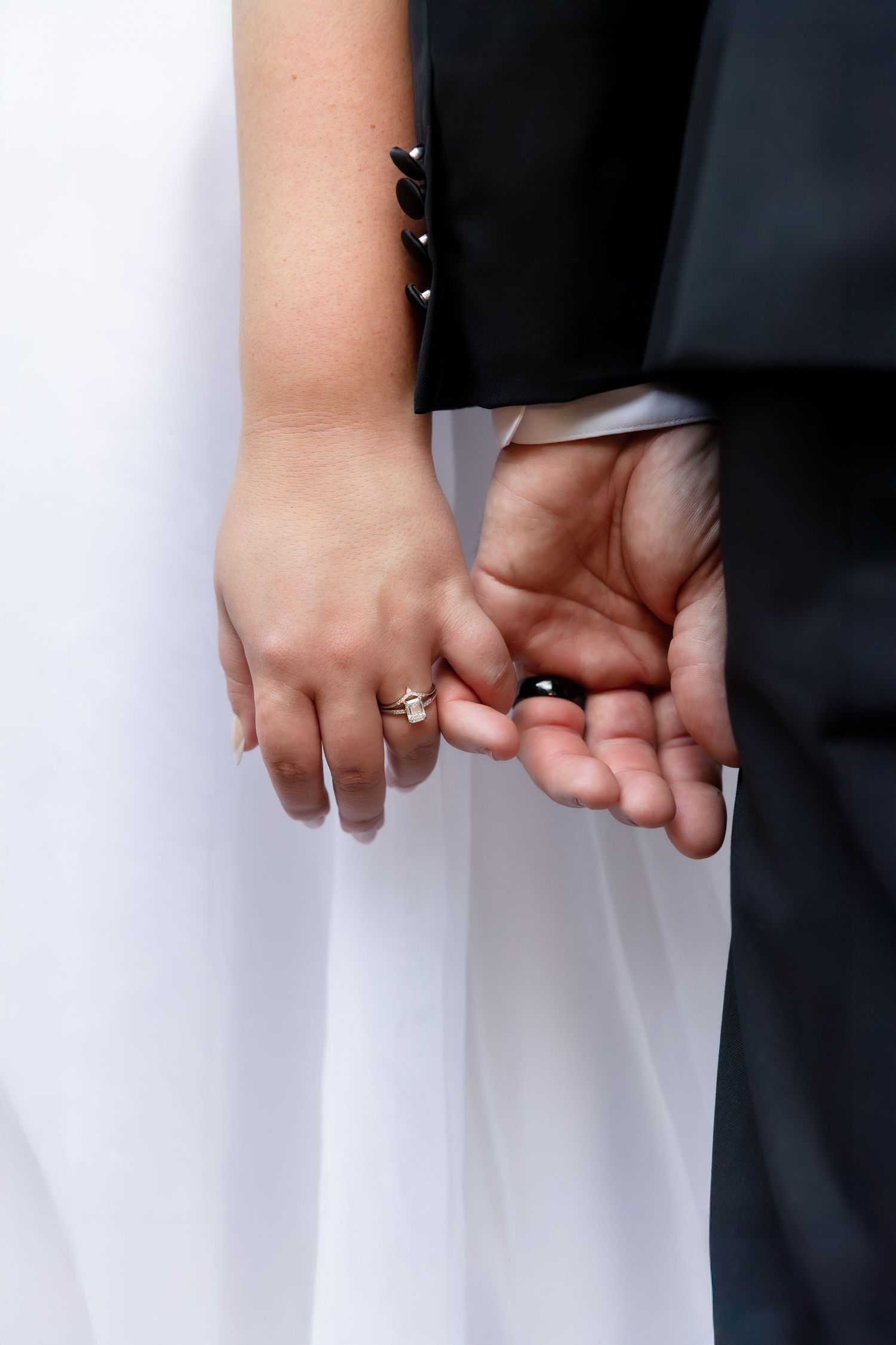Close-up view of hands holding together showing wedding rings against black and white formal attire.