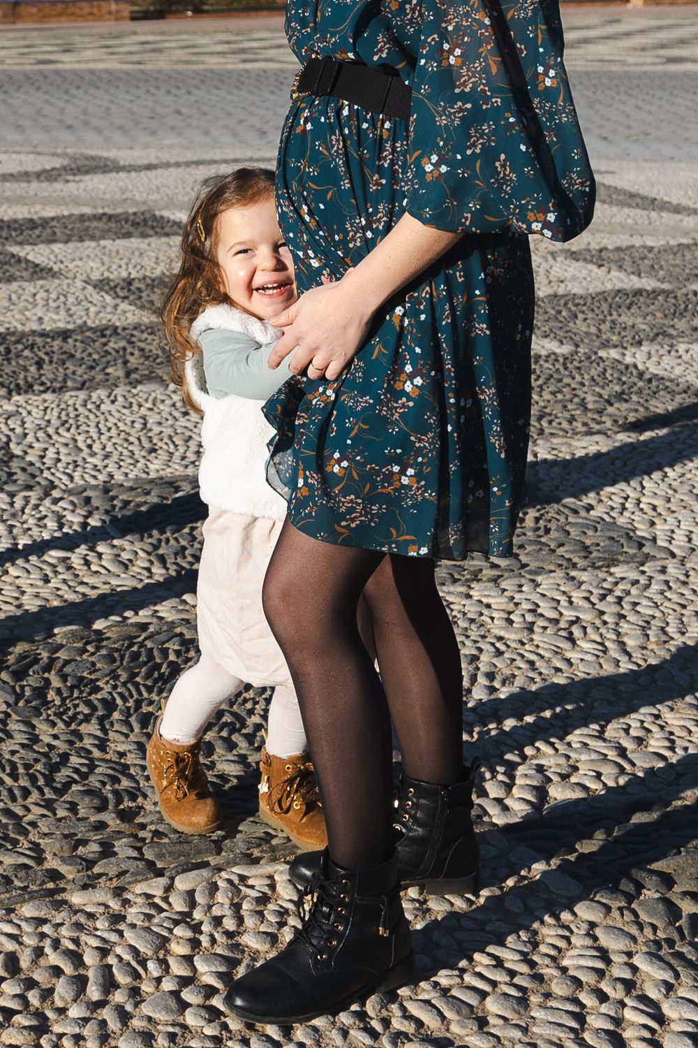 A small child in white clothing hugs someone wearing a floral blue dress and black tights on a cobblestone street.