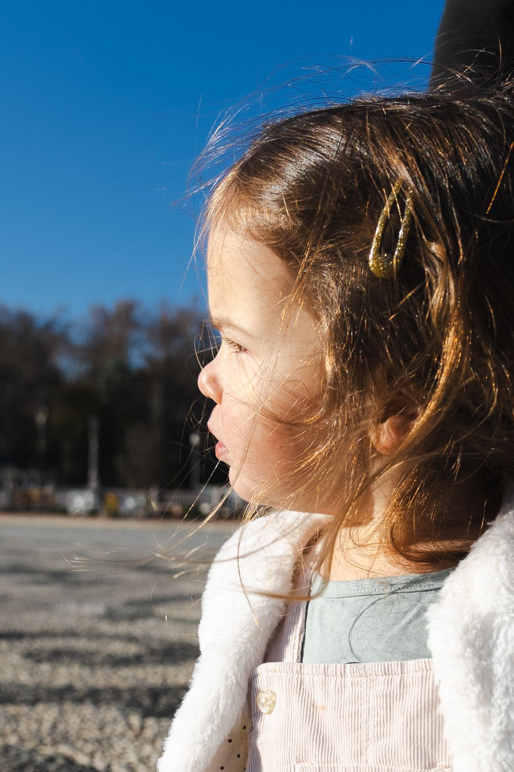A child in a white sweater and overalls gazes into the distance on a sunny day with wispy hair blowing in the breeze.