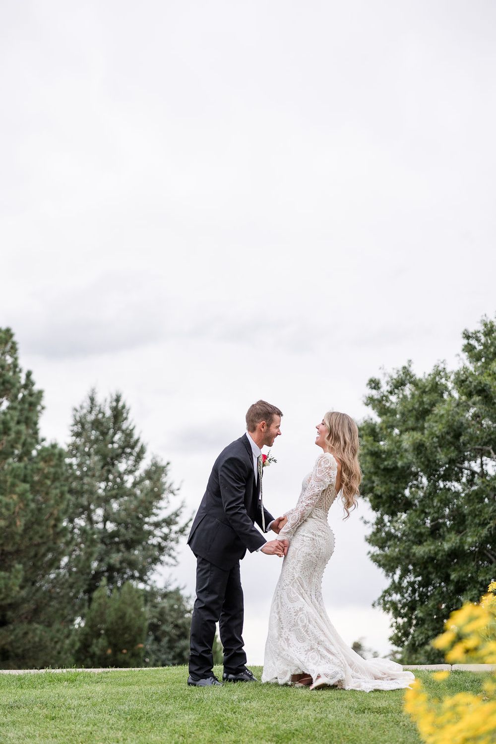 A couple in wedding attire playfully interacts on a grassy hillside with trees in the background.