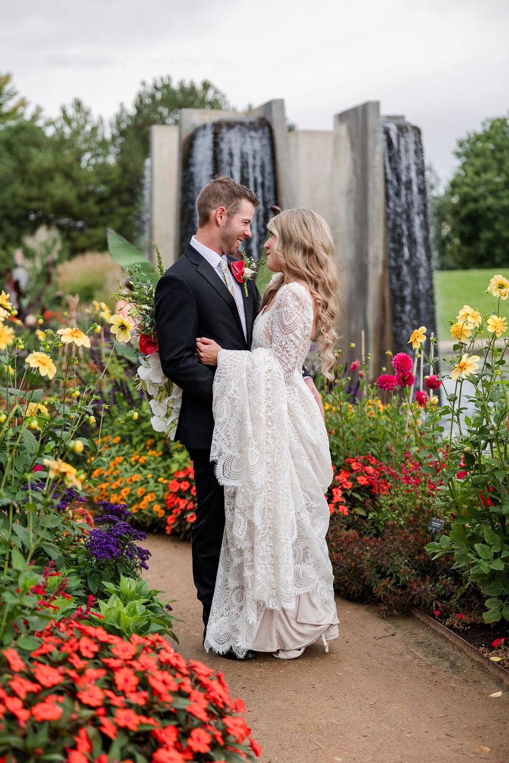 A couple in wedding attire poses in a garden with colorful flowers and stone pillars at sunset.