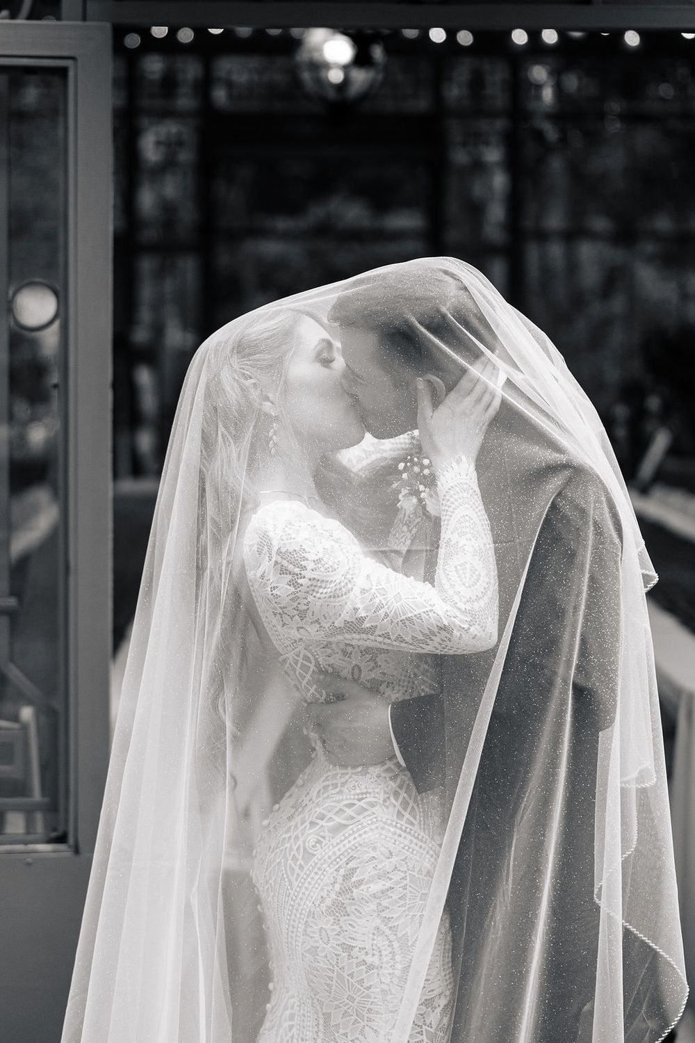 A romantic black and white wedding photograph showing a couple sharing a tender moment under a flowing veil.
