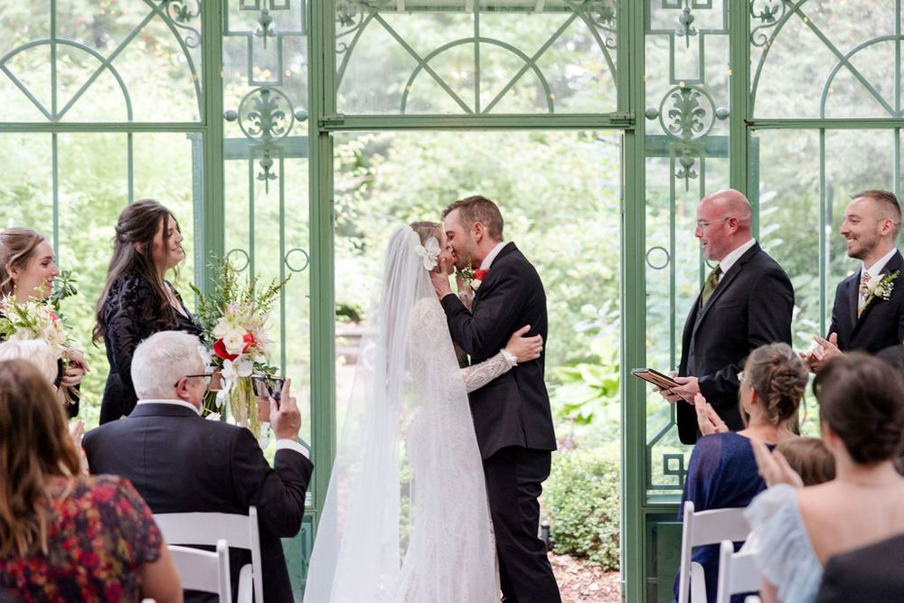 Wedding ceremony takes place in an elegant glass conservatory filled with natural light and greenery.