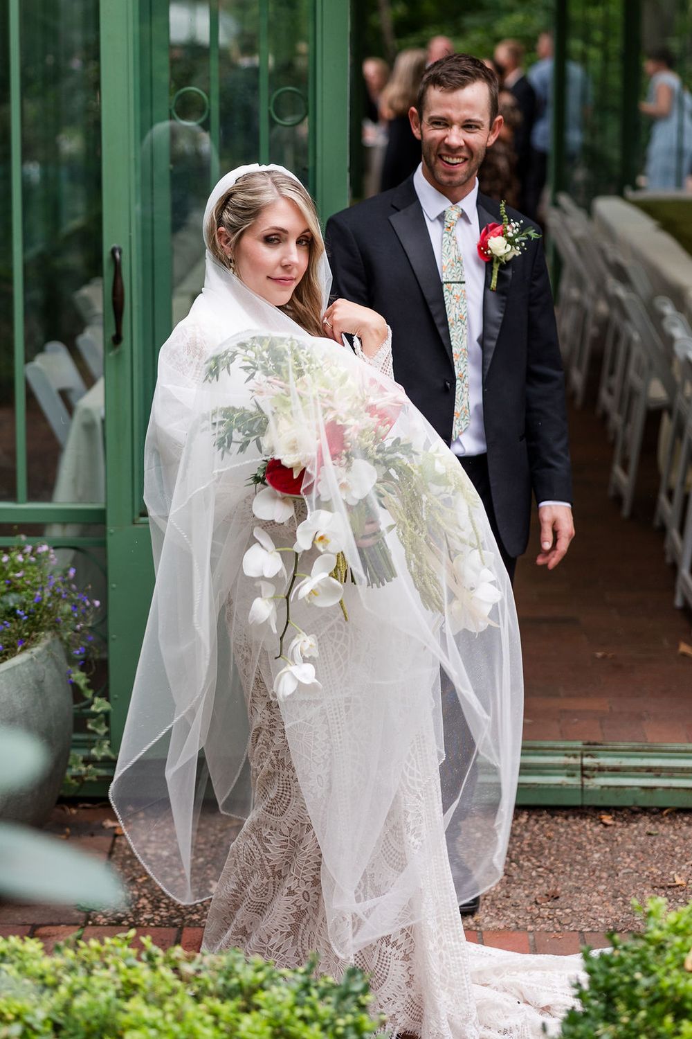 A bride in a white lace dress and veil walks along a garden path with ornate green architecture in the background.