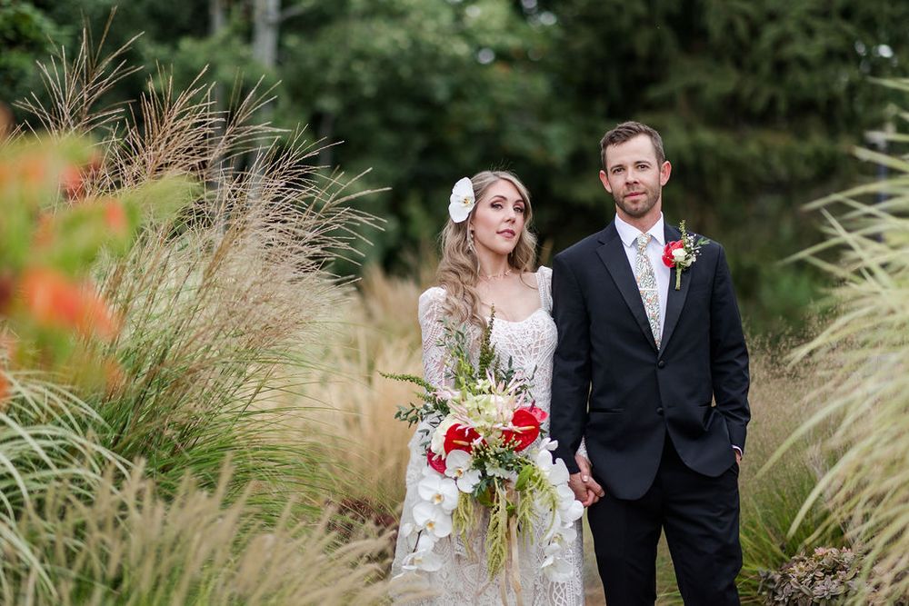A couple in wedding attire poses together in a garden setting with ornamental grasses and autumn foliage.