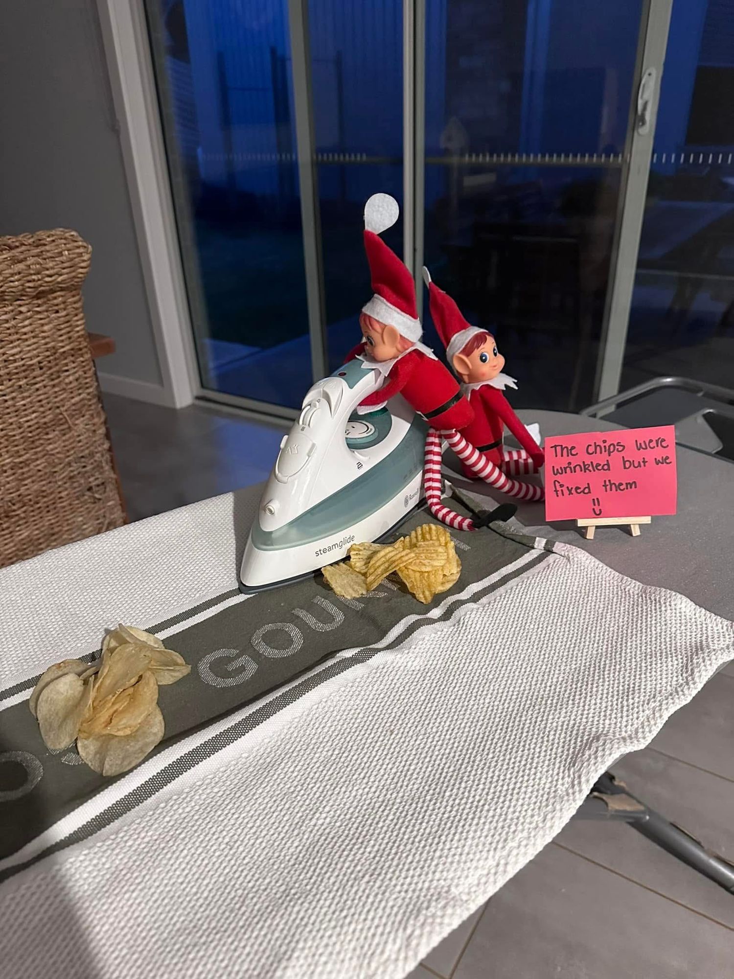 A holiday elf doll sits playfully on an iron while positioned on a folded gray cloth with a handwritten note nearby.