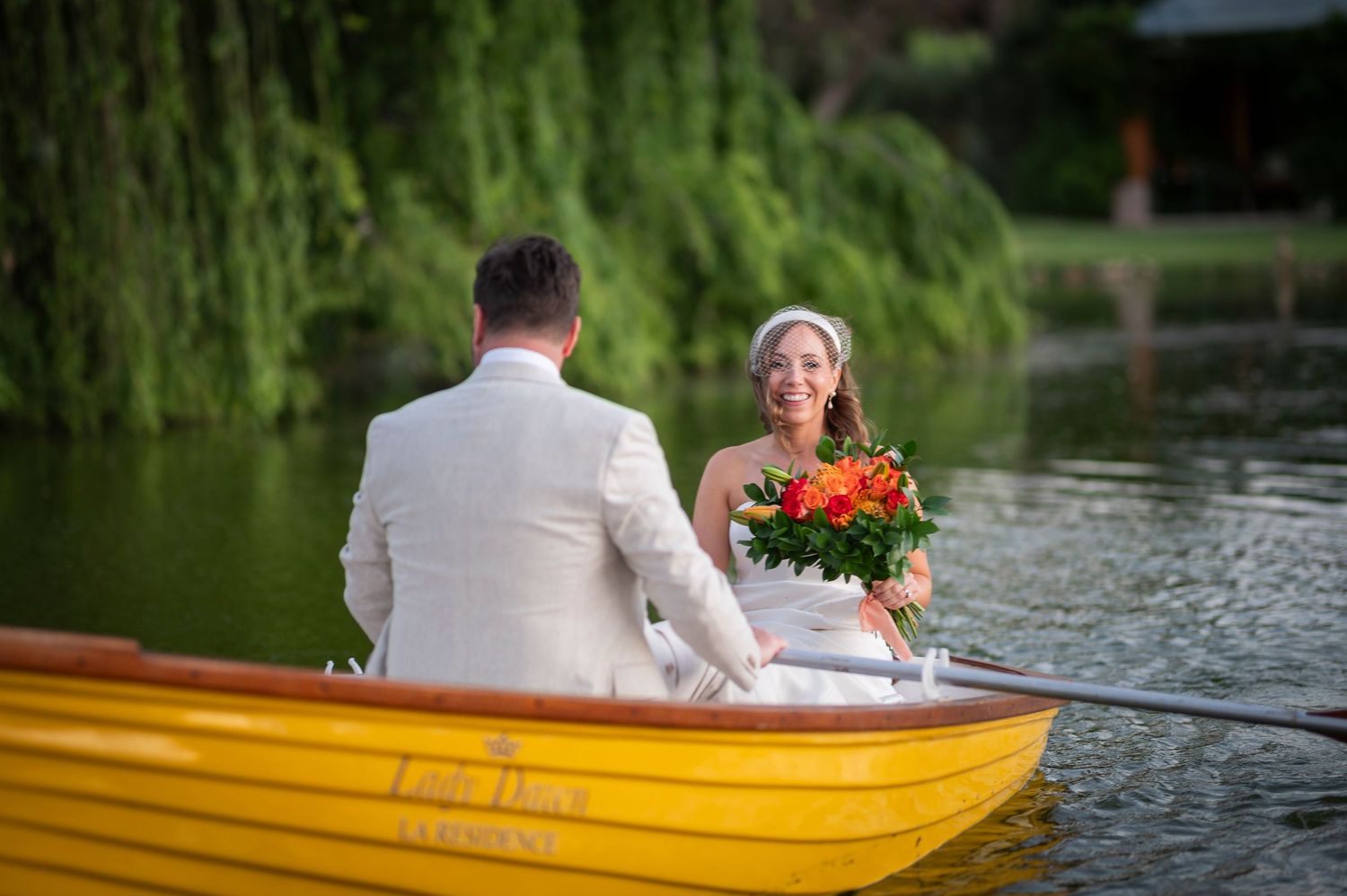 A couple in wedding attire sits in a yellow rowboat on a peaceful lake with weeping willows in the background.
