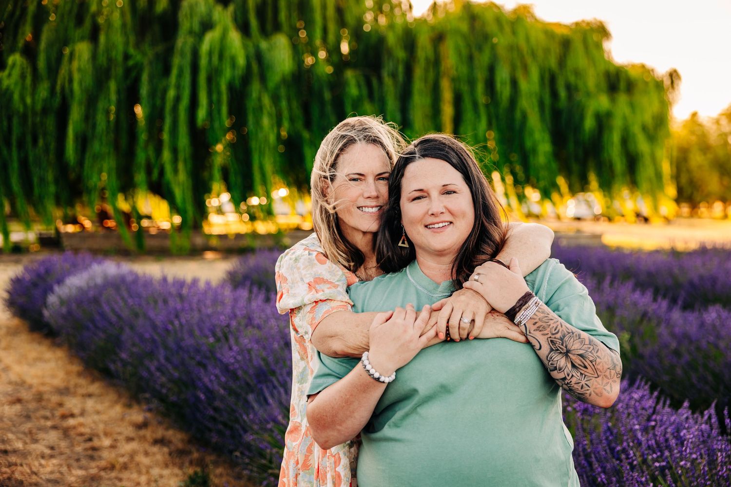 Two people embrace in a lavender field at sunset with golden light filtering through willow trees in the background.