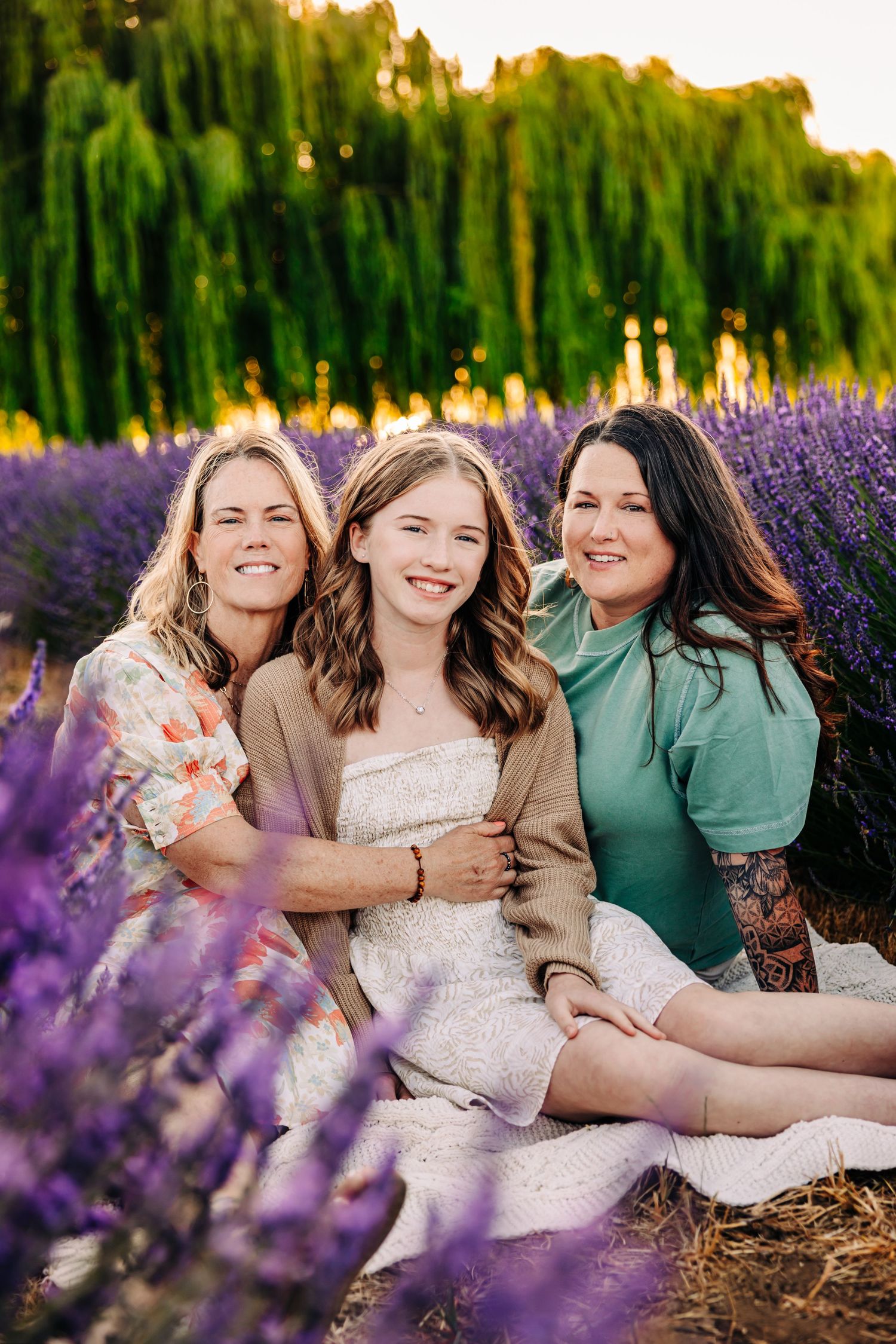 Three friends share joyful laughter while sitting together in a blooming lavender field at sunset with willow trees behind them.
