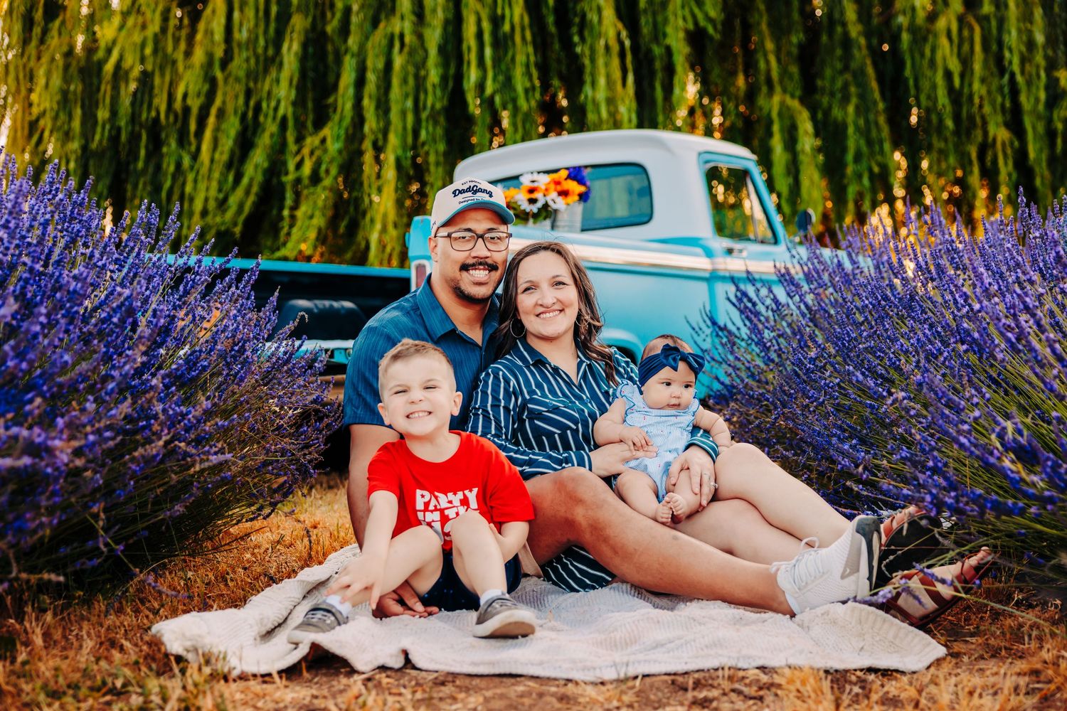 A group sits on a blanket beside blooming lavender and a vintage blue pickup truck in a garden setting.