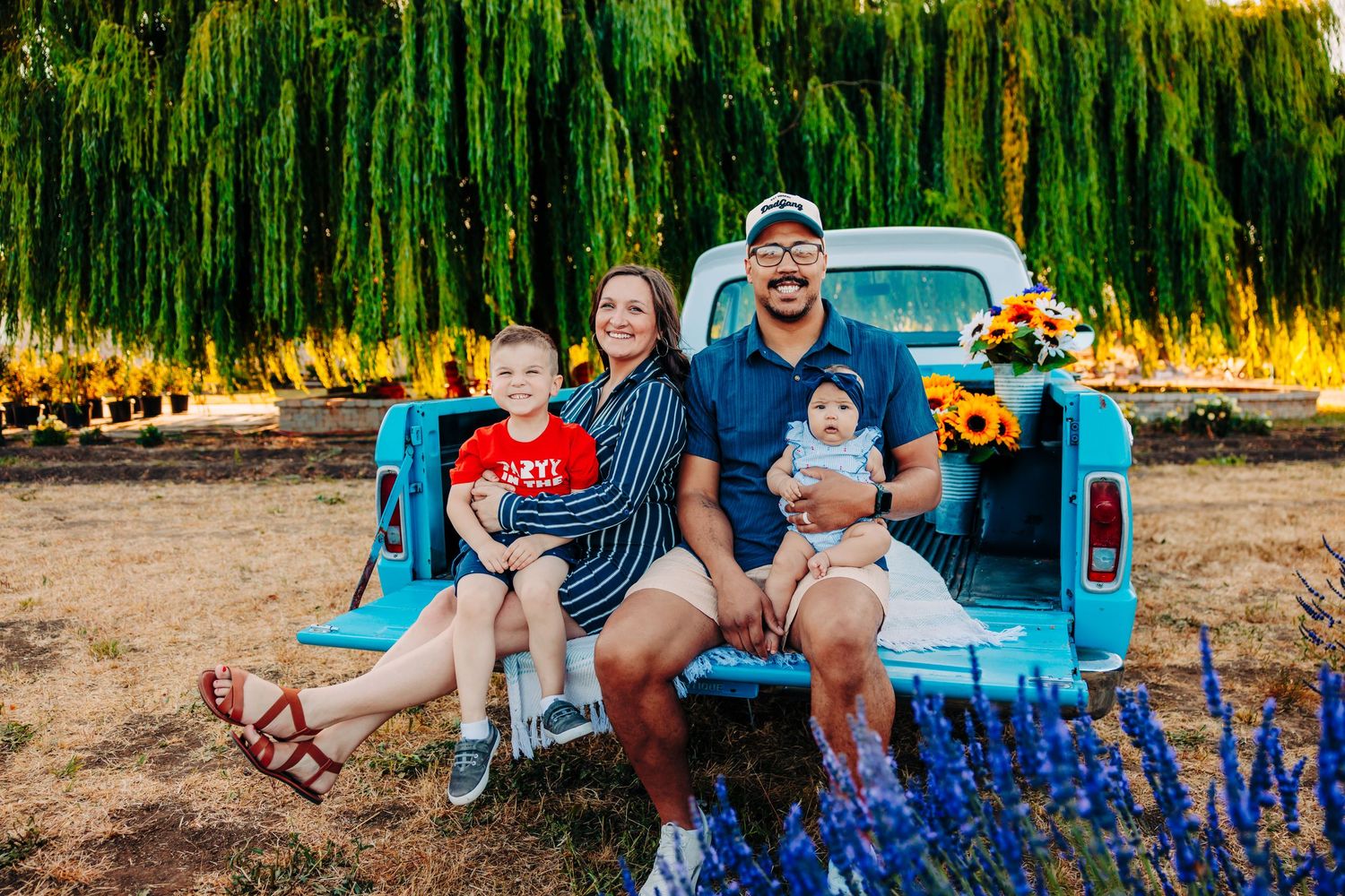 A family sits on the back of a vintage blue pickup truck surrounded by vibrant wildflowers in a sunny field.