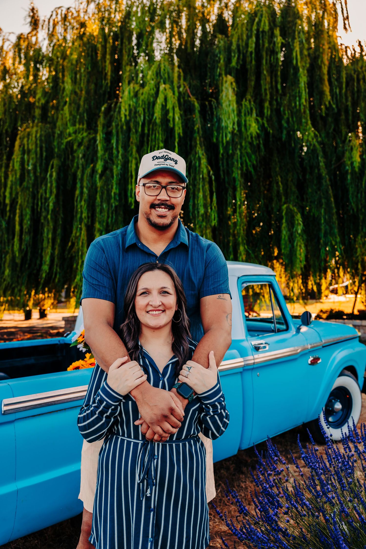 A couple poses together in front of a vintage blue car and blooming lavender field at sunset.