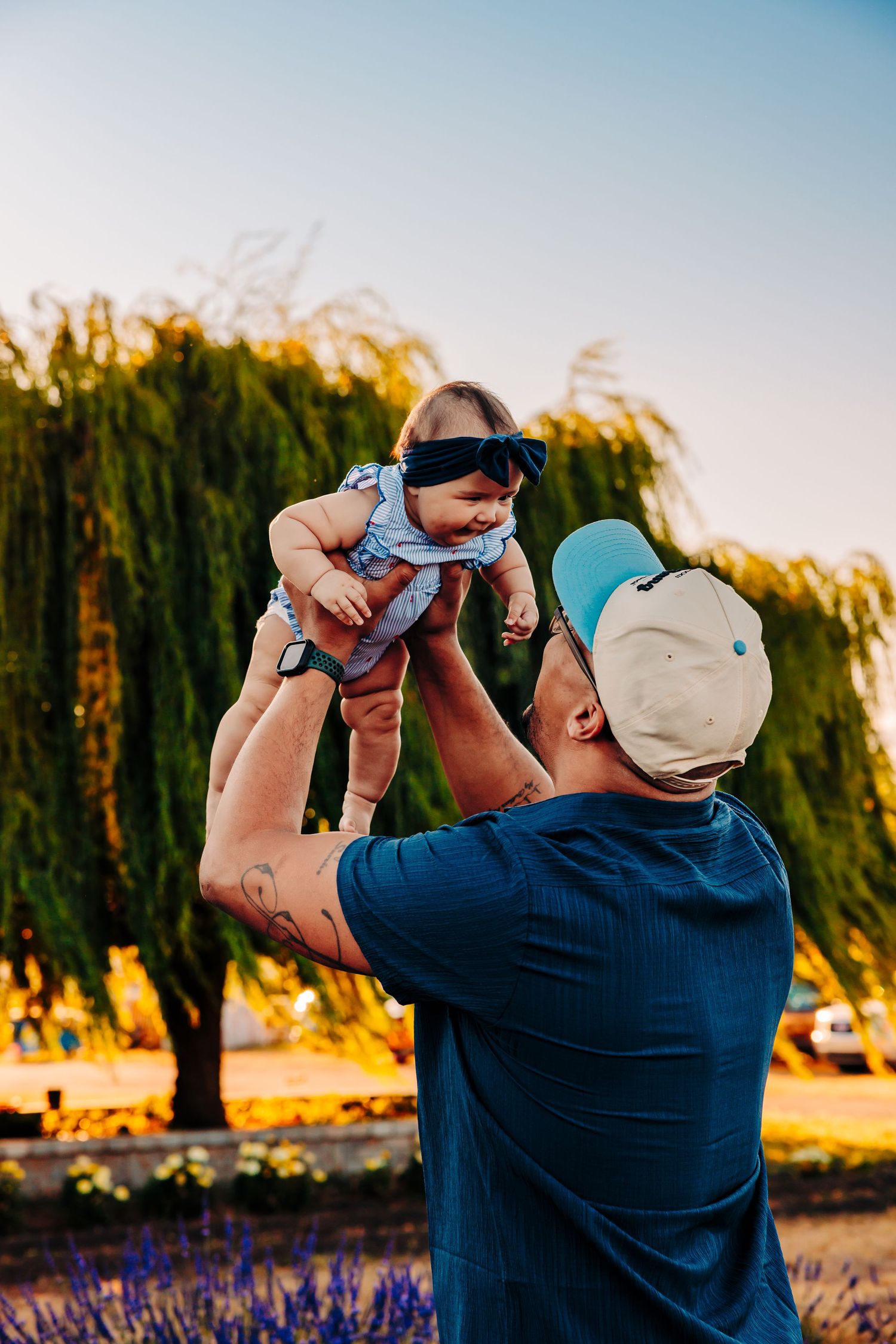 A parent lifts a baby up in the air during golden hour with willow trees in the background at sunset.