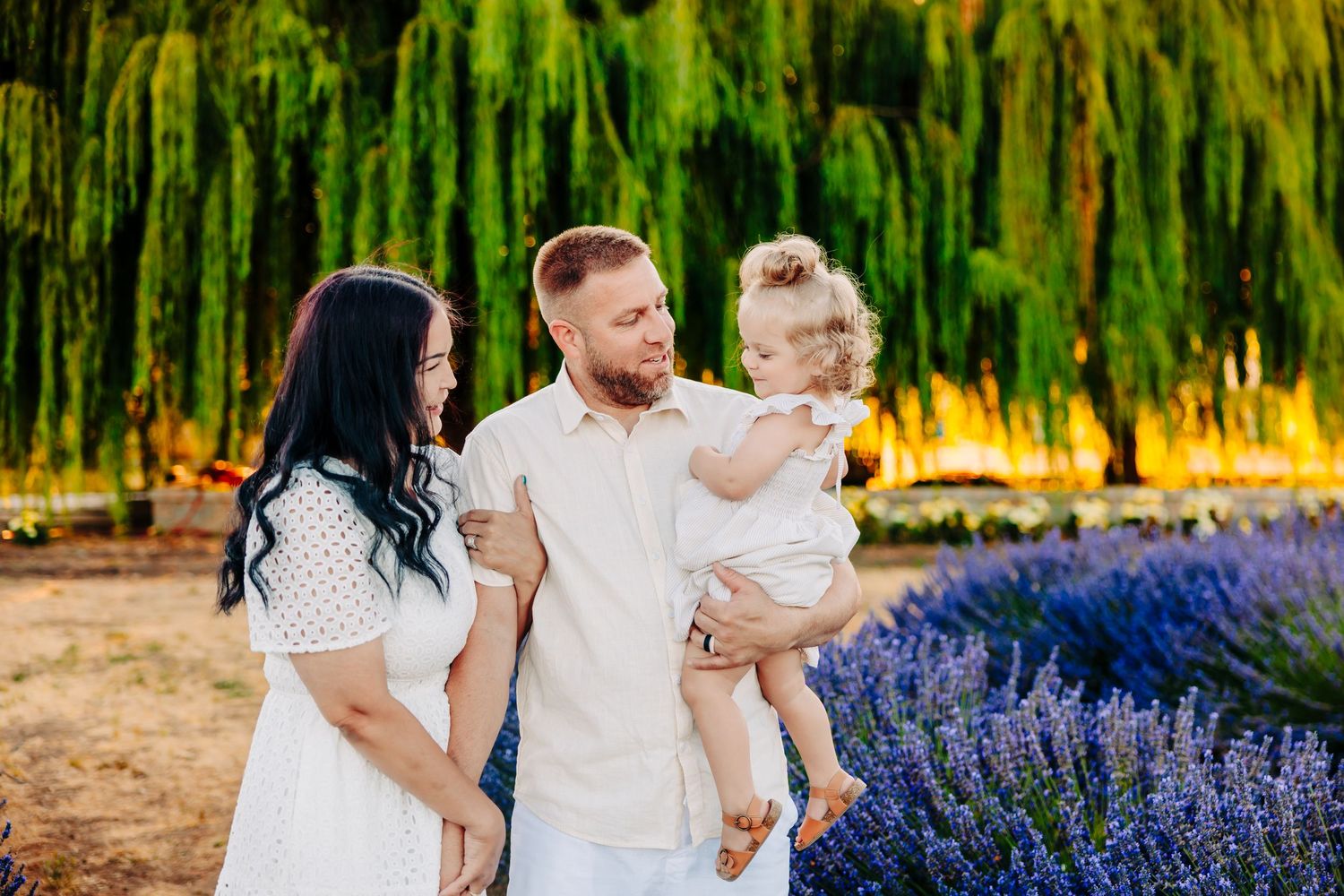 Family wearing white outfits poses together in a garden with blooming purple lavender and weeping willows in background.