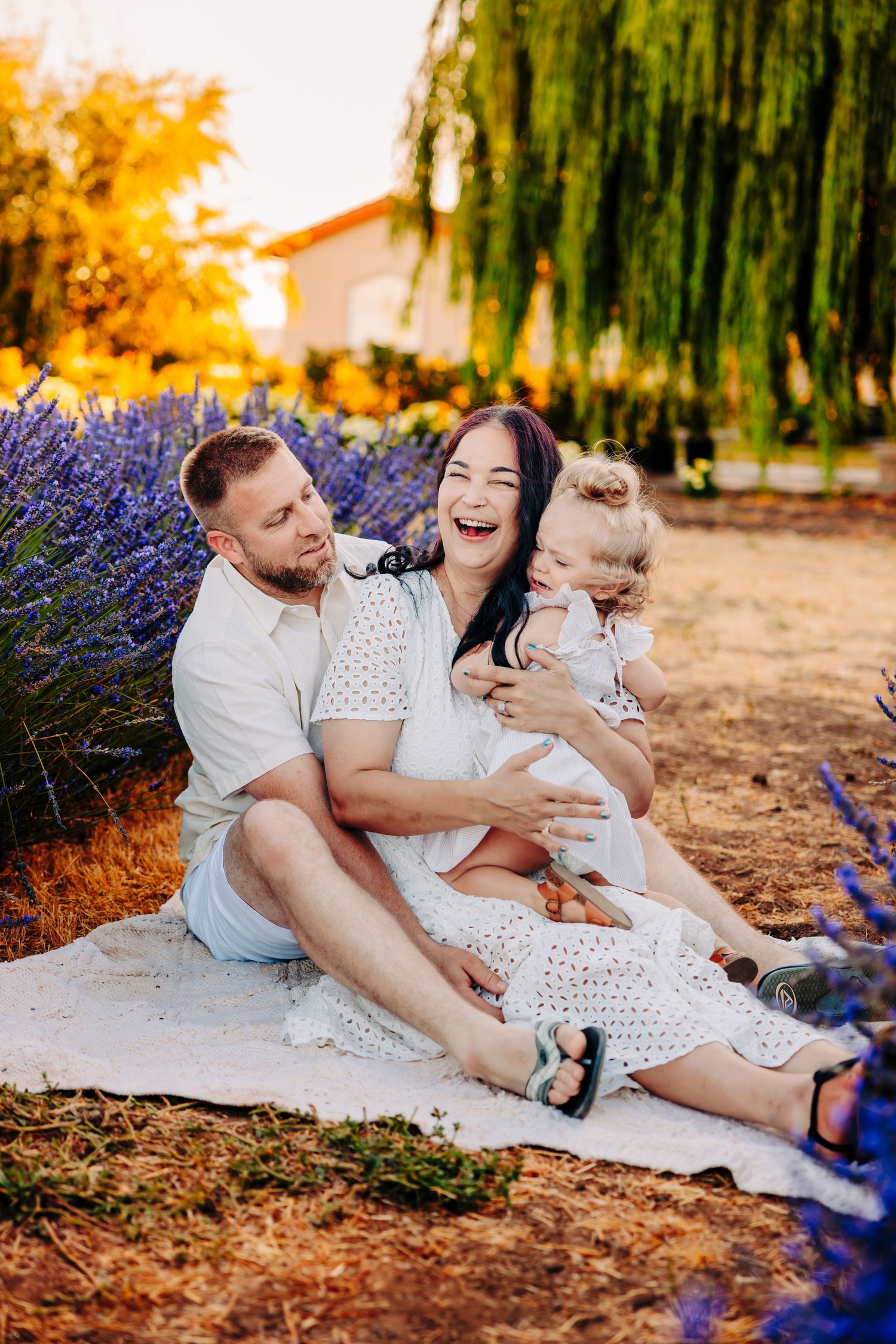 A family relaxes together on a blanket at sunset with willow trees and purple flowers in the background.