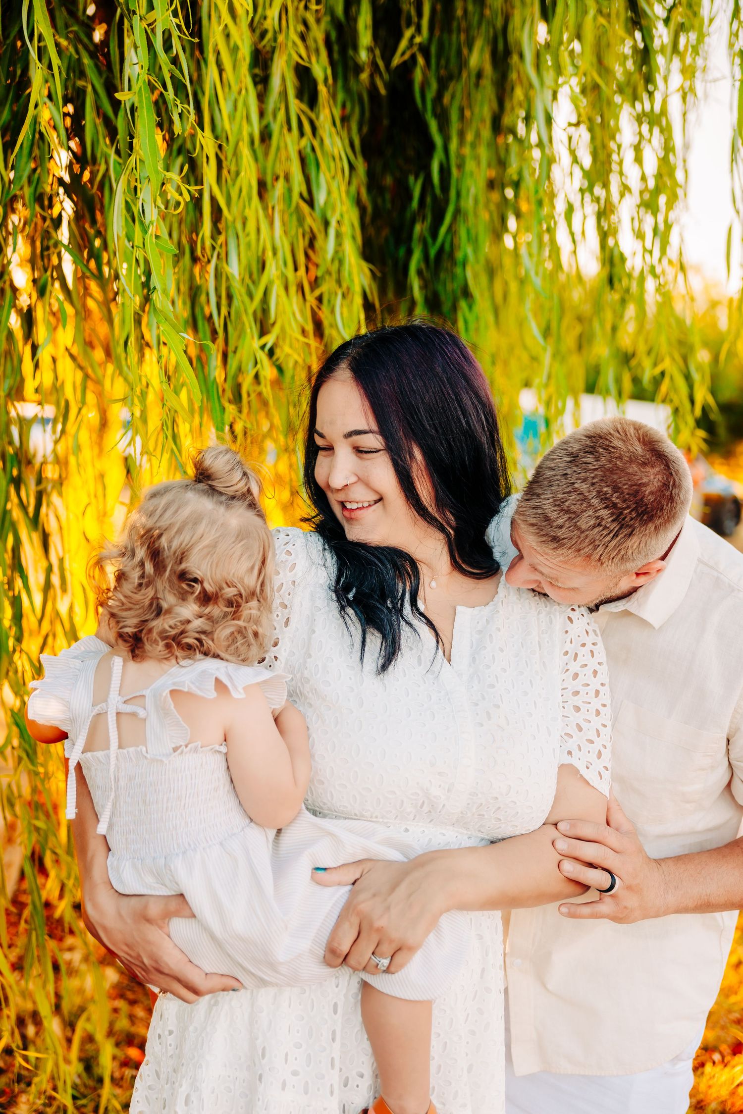 Family in white outfits embraces and laughs together under golden willow tree branches during sunset photoshoot.