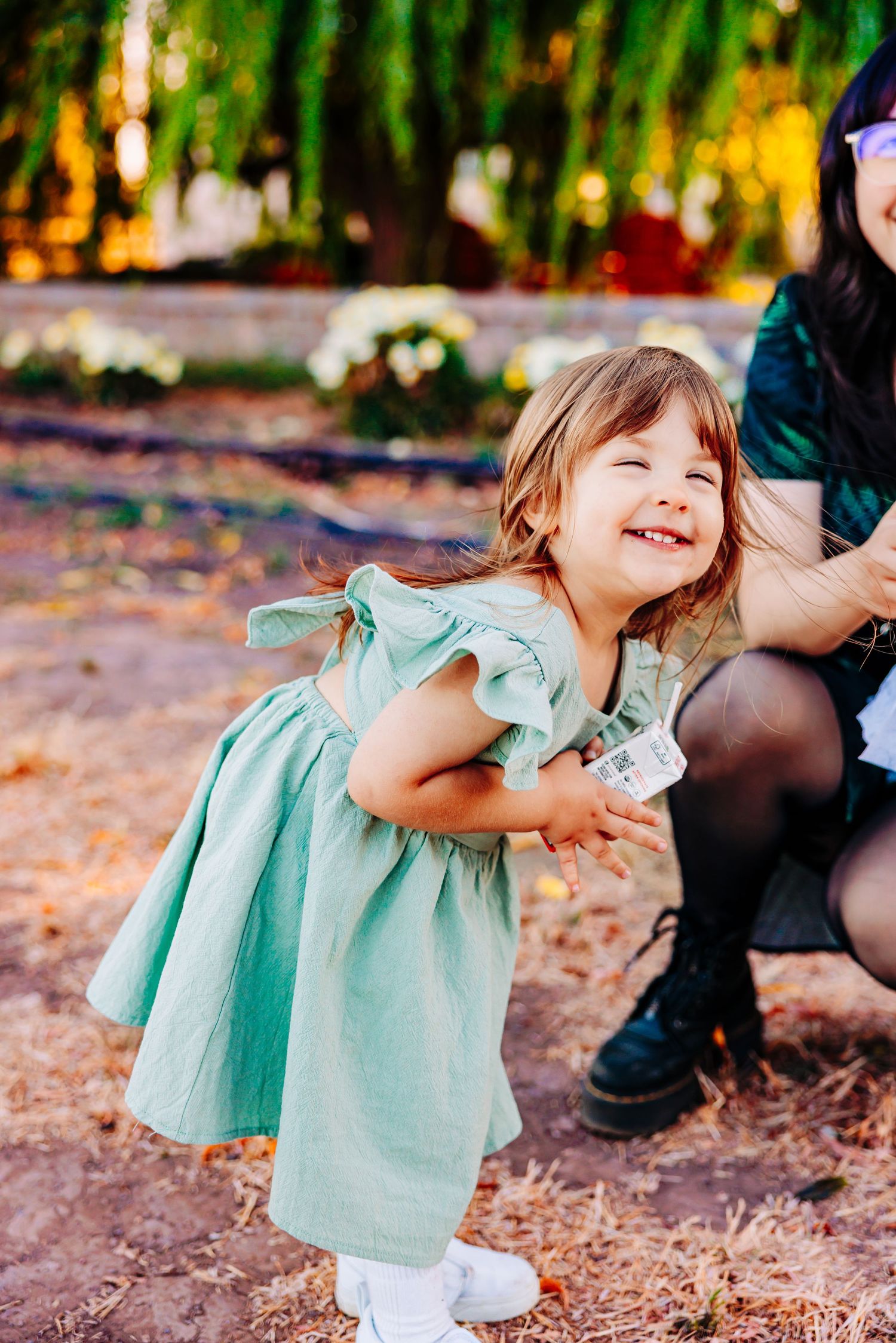 A child in a mint green dress twirls playfully in a park setting with autumn leaves on the ground.