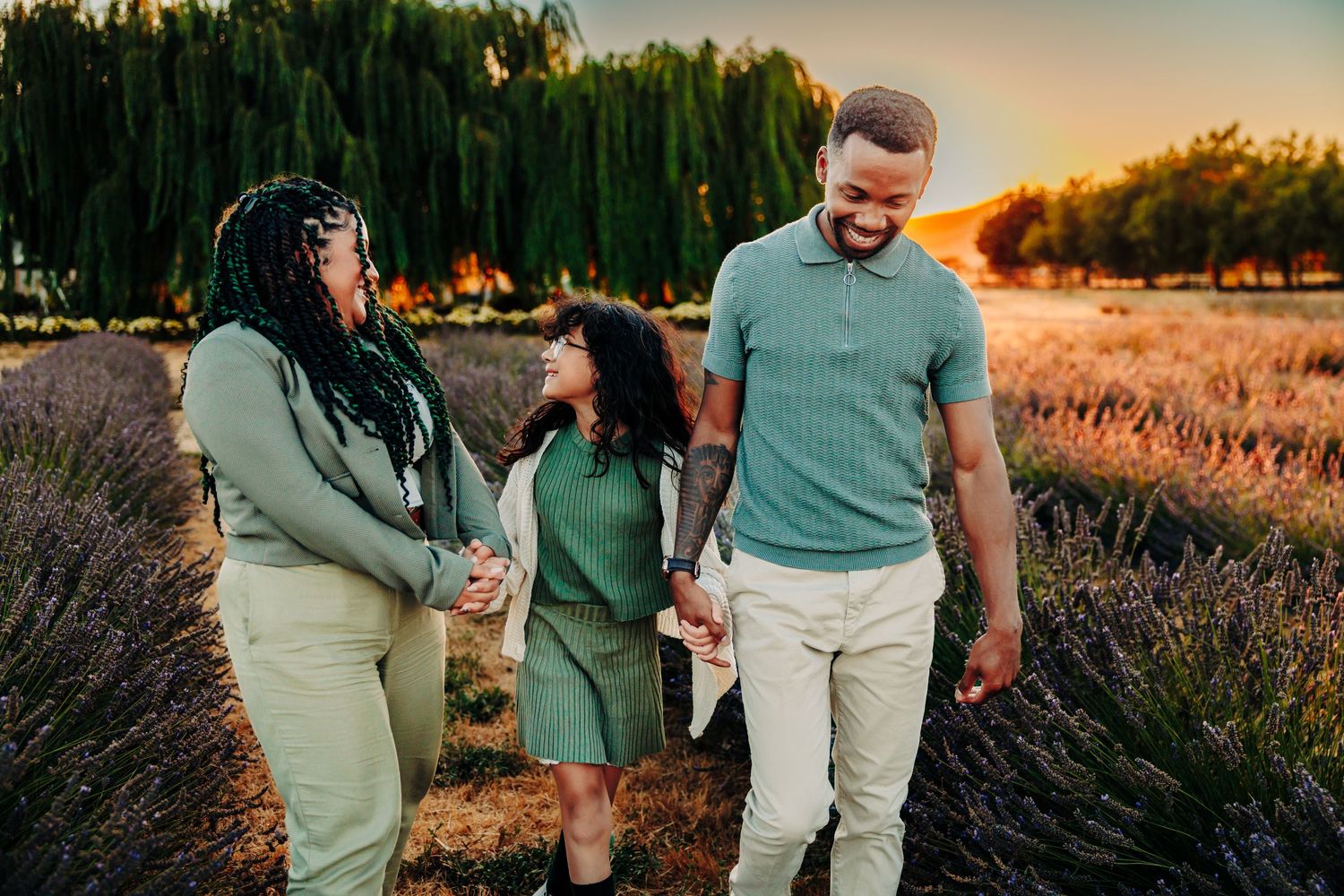 Family portrait session in a lavender field at golden hour with warm sunset lighting and willow trees in background.