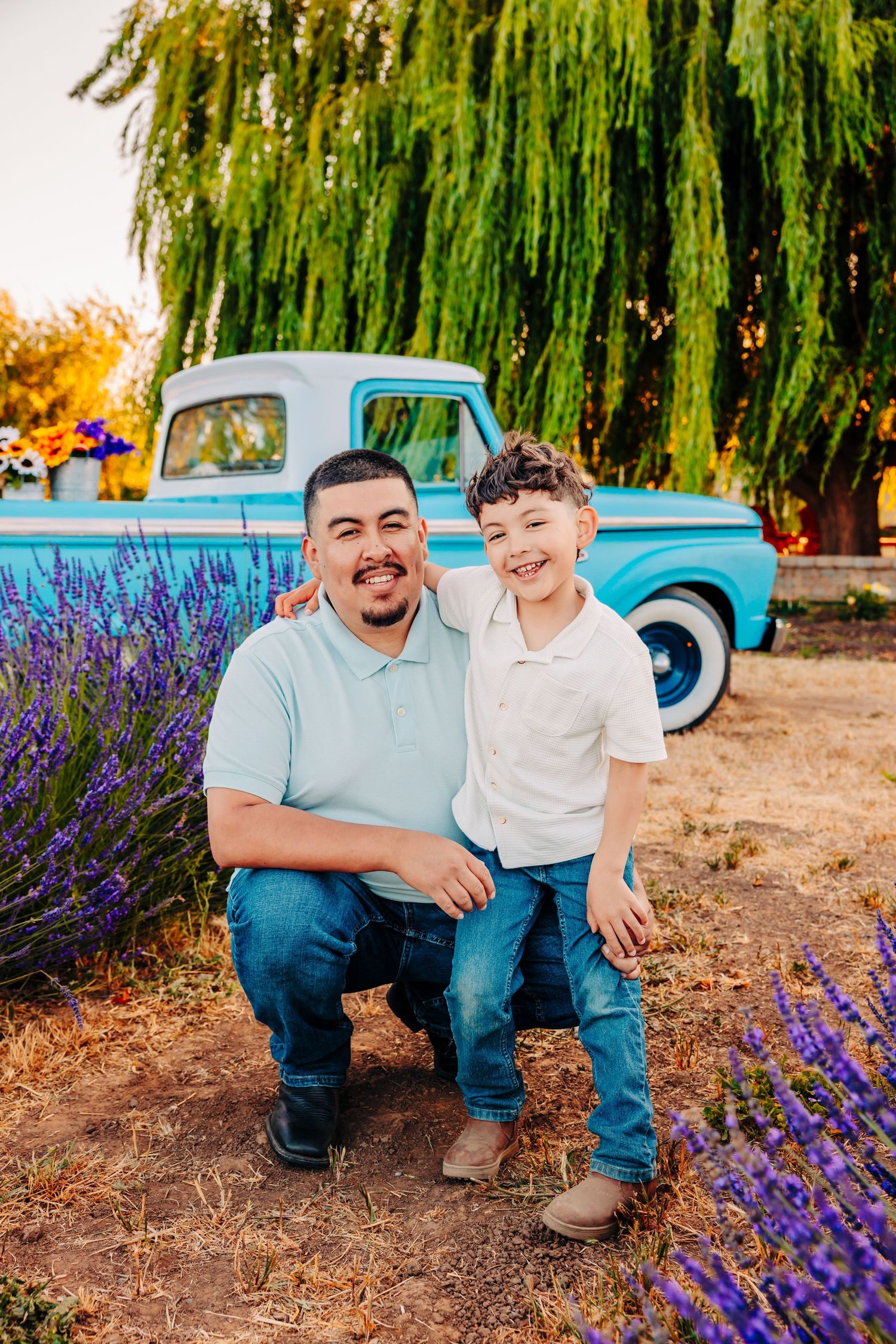 A vintage blue pickup truck serves as backdrop for family photos among blooming purple lavender and willow trees.