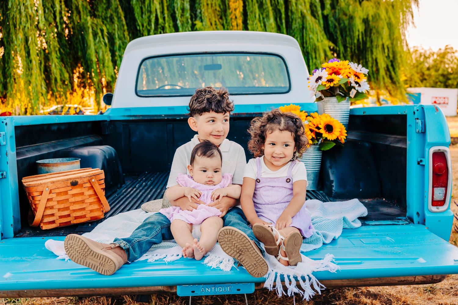 A vintage turquoise pickup truck bed filled with pillows and blankets creates a cozy outdoor seating area at sunset.