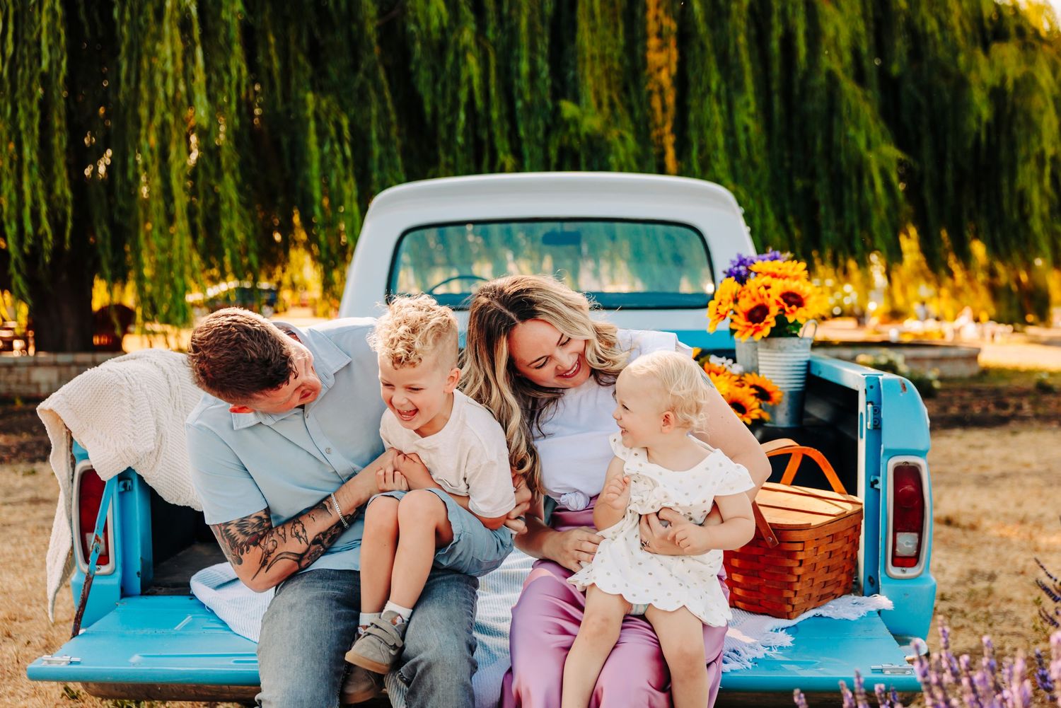 A family enjoys quality time together while sitting in the bed of a vintage blue pickup truck during golden hour.