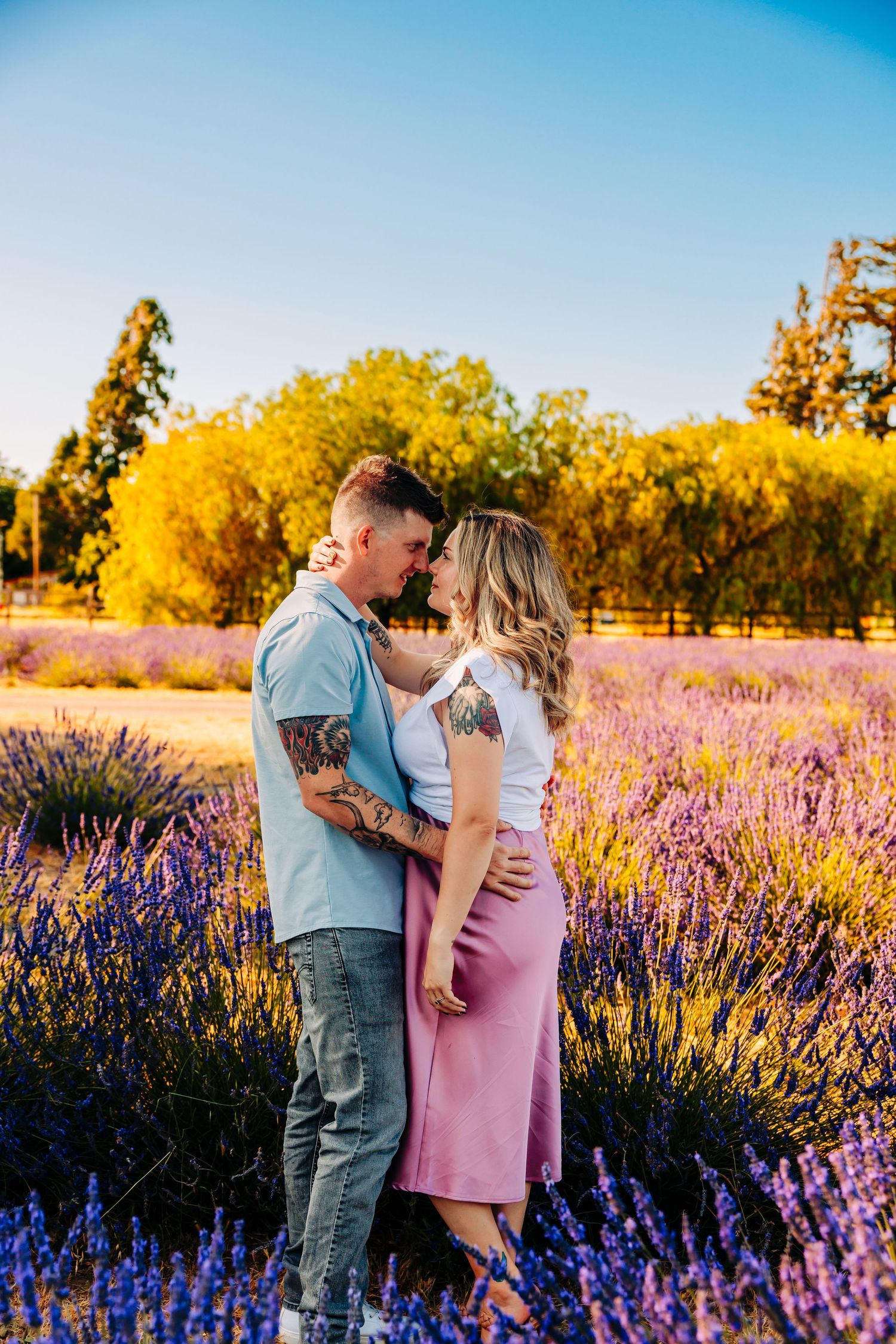 A romantic couple embraces in a blooming lavender field with golden trees and blue sky in the background.