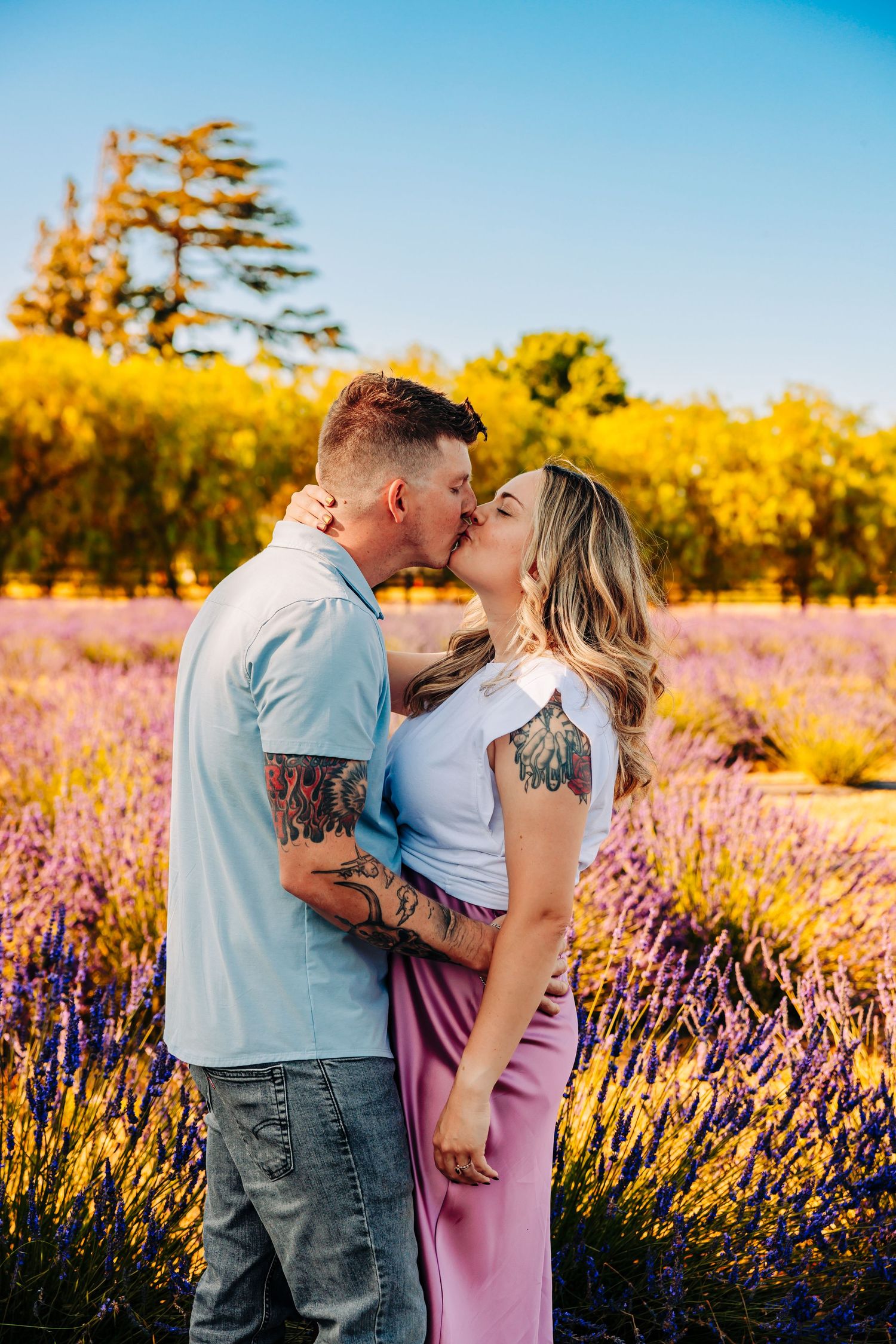 A couple shares a romantic moment in a lavender field during golden hour with autumn trees in the background.