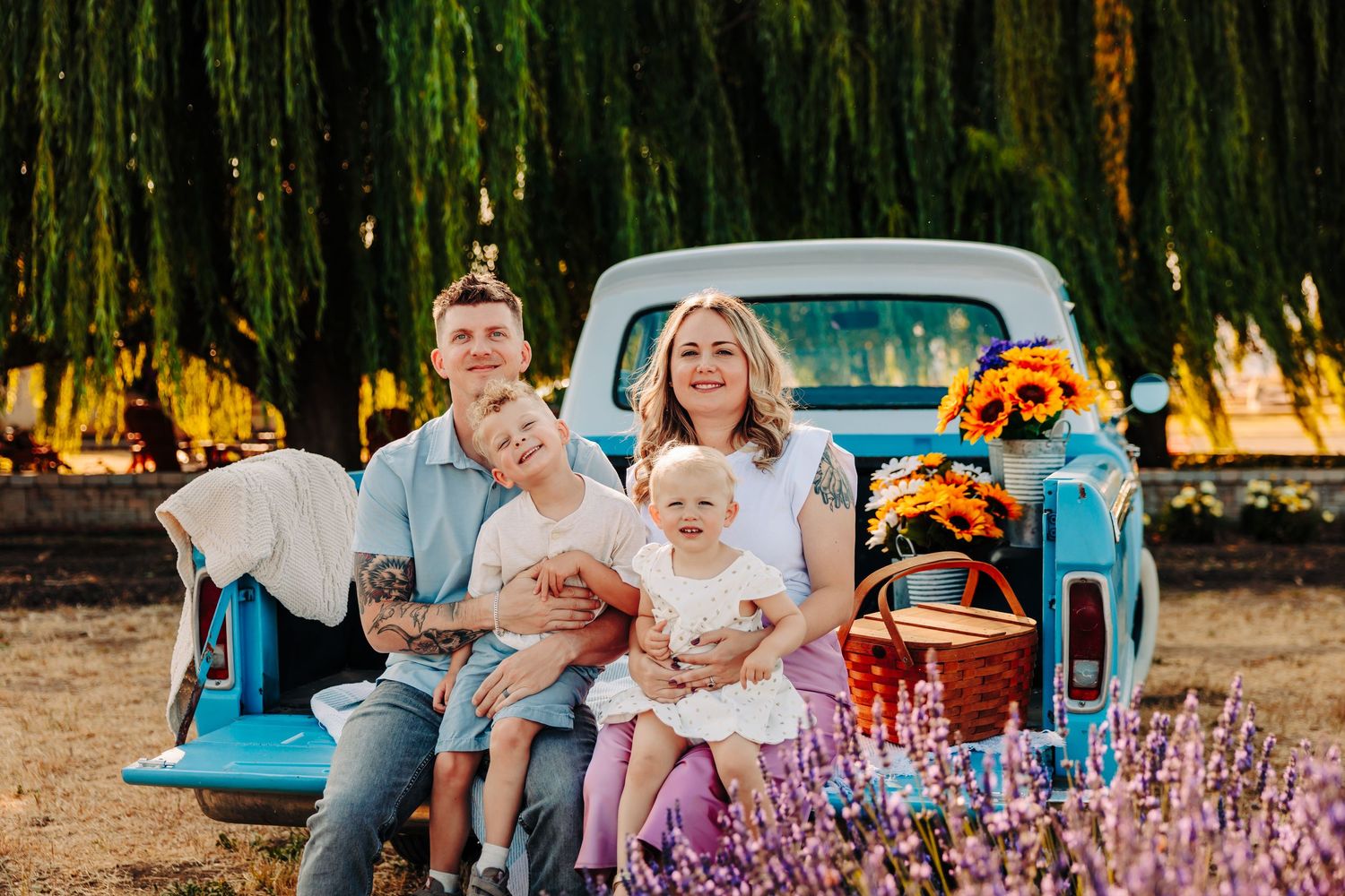 Family sits together on vintage blue pickup truck with sunflowers at sunset in rustic outdoor setting.