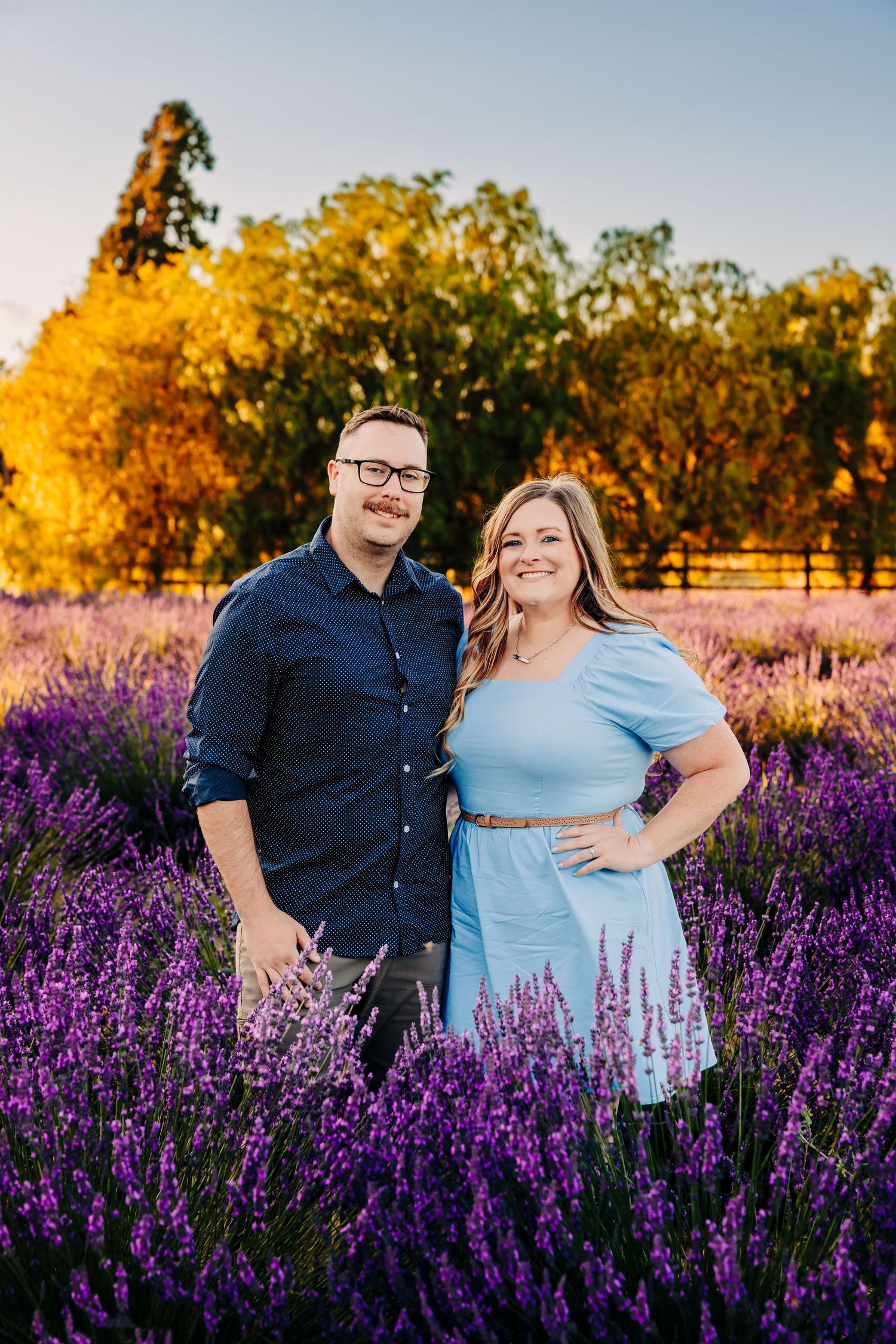 A couple poses together in a vibrant field of purple lavender and golden autumn trees during sunset.