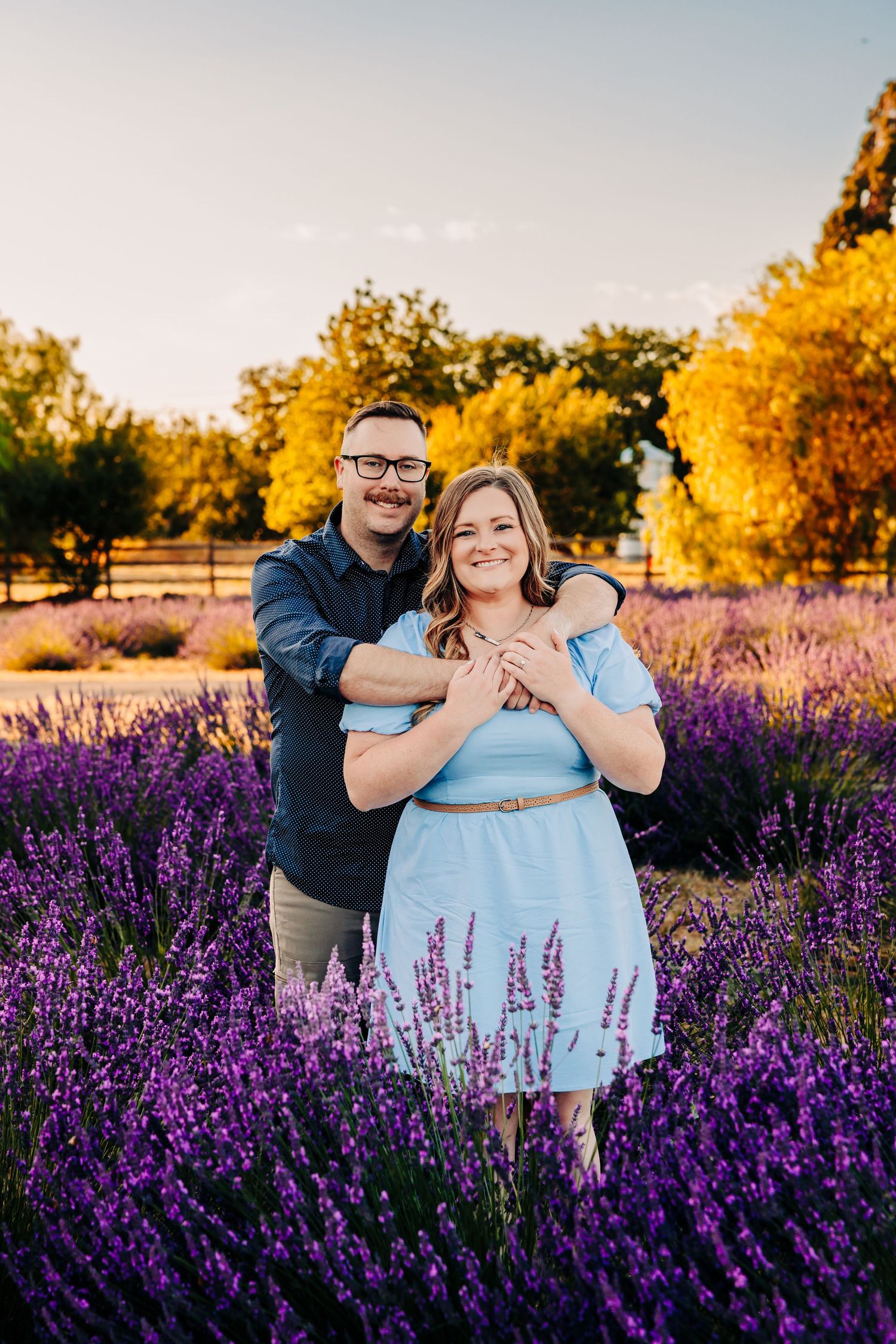 A couple embraces in a vibrant lavender field at golden hour with autumn trees in the background.