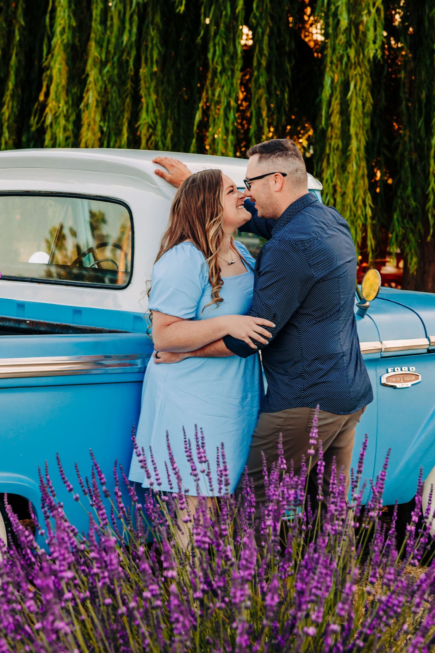 A couple shares a romantic moment next to a vintage blue car with purple lavender flowers blooming in the foreground.