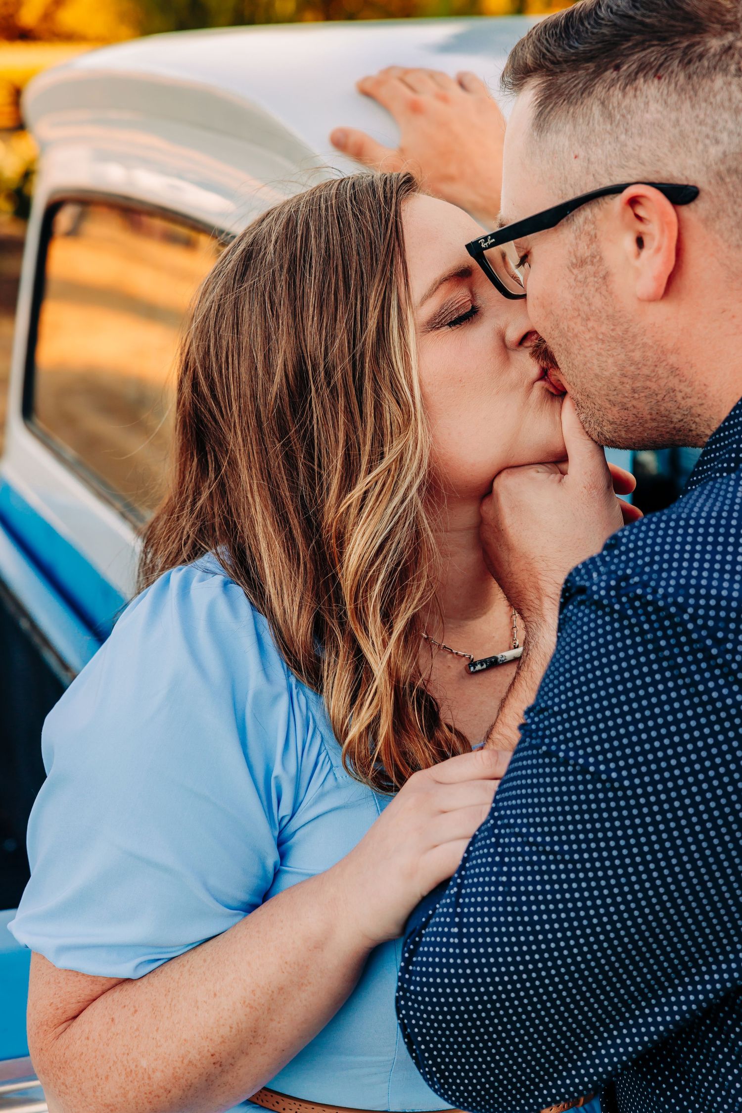 A couple shares a romantic kiss while leaning against a vintage car during a golden sunset.