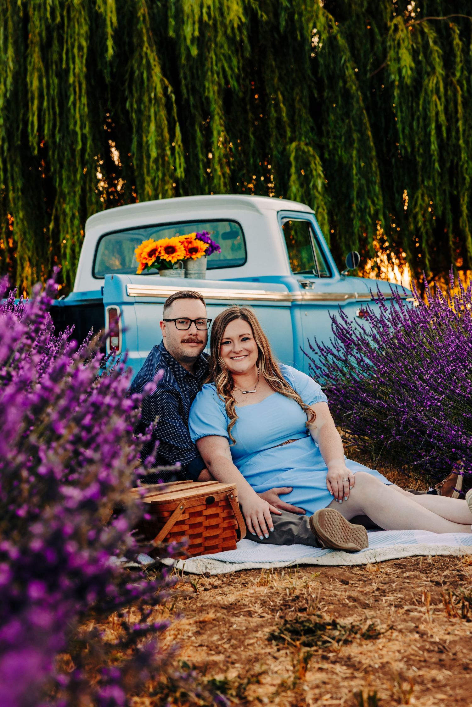A couple relaxes on a blanket near vibrant purple lavender flowers and weeping willow trees during a summer photoshoot.