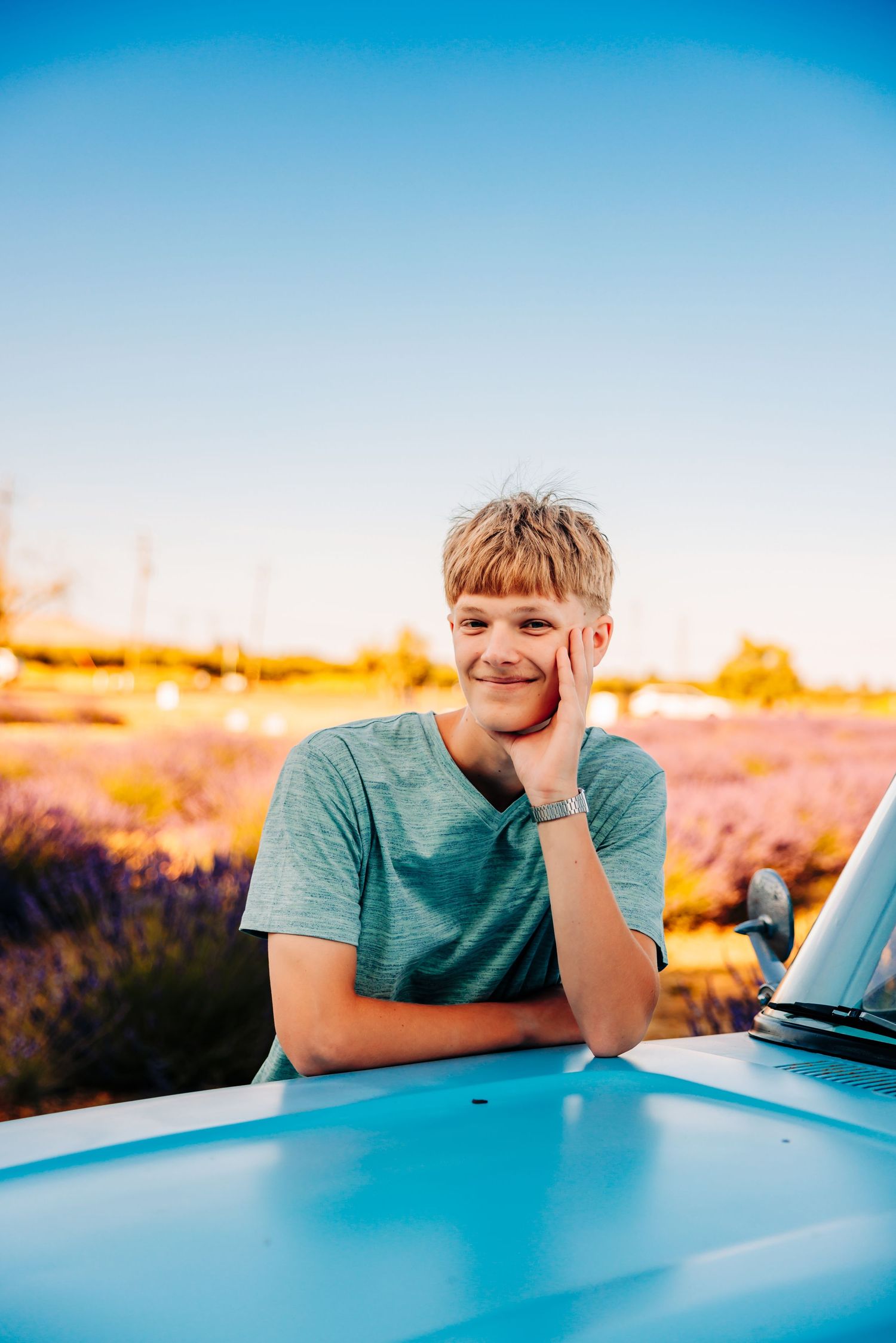 A person in a teal shirt leans on a car during golden hour sunset, creating a serene outdoor portrait series.